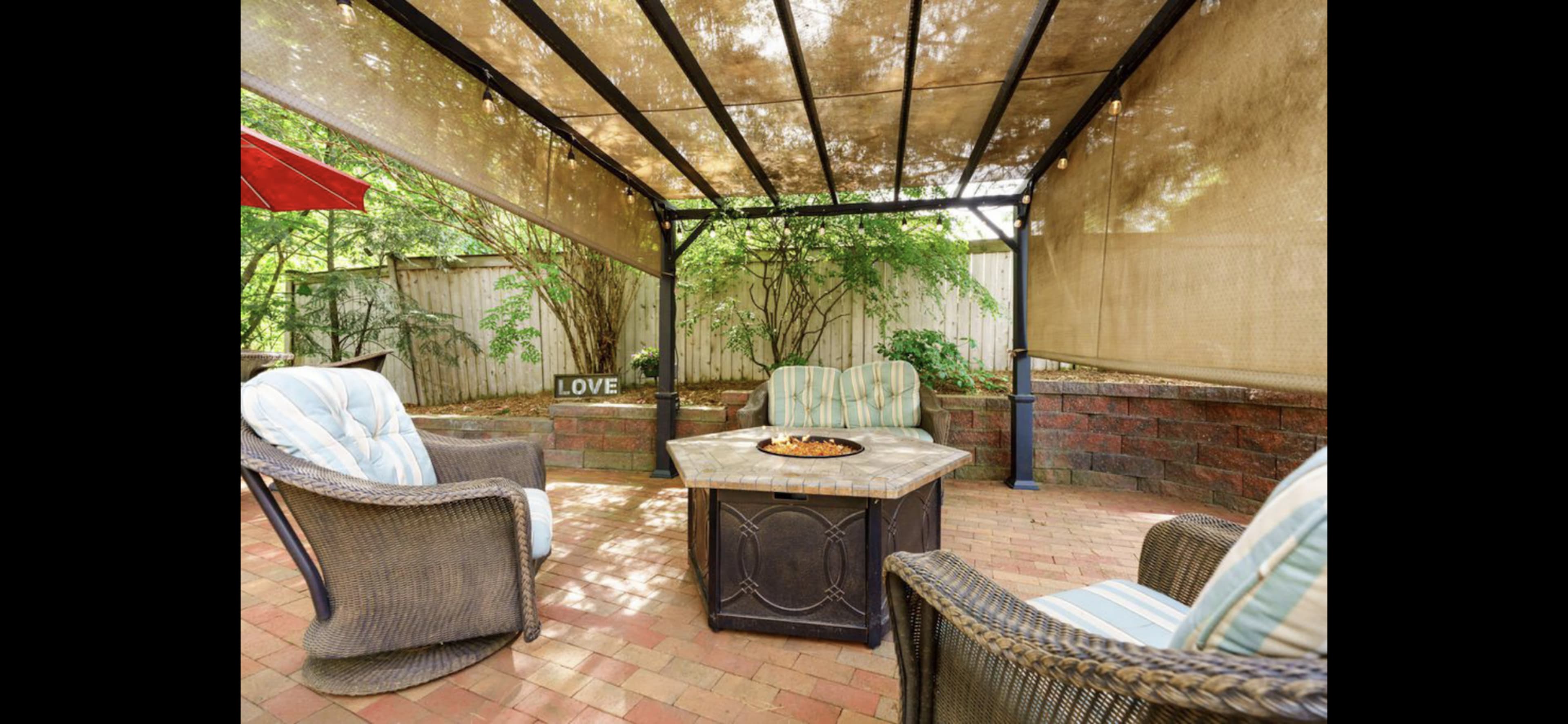 A covered patio features four wicker chairs surrounding a fire pit, set against a backdrop of greenery and a decorative "LOVE" sign.