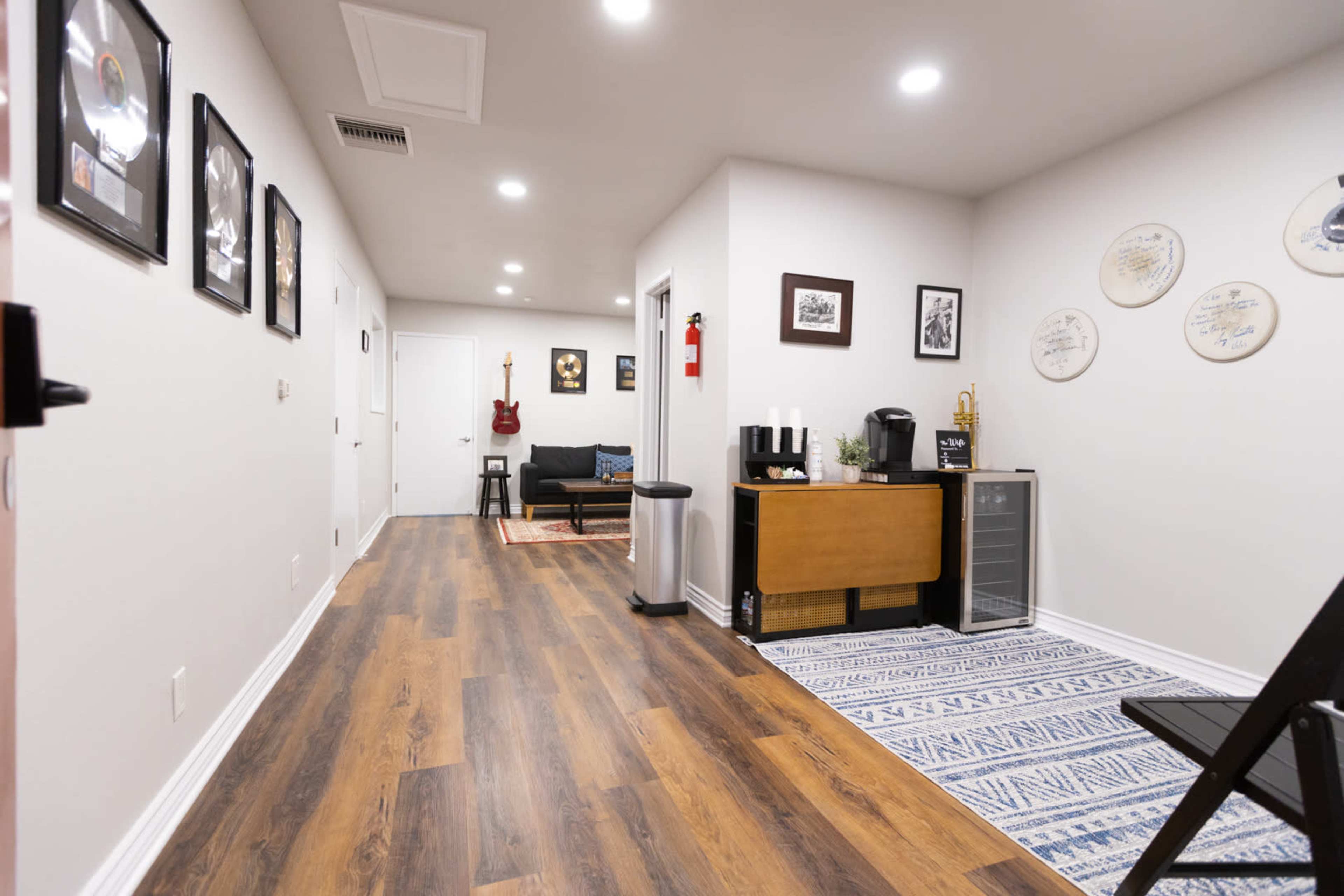 The image shows a long, well-lit hallway leading to a living area with a couch, a small table, and framed artwork on the walls.