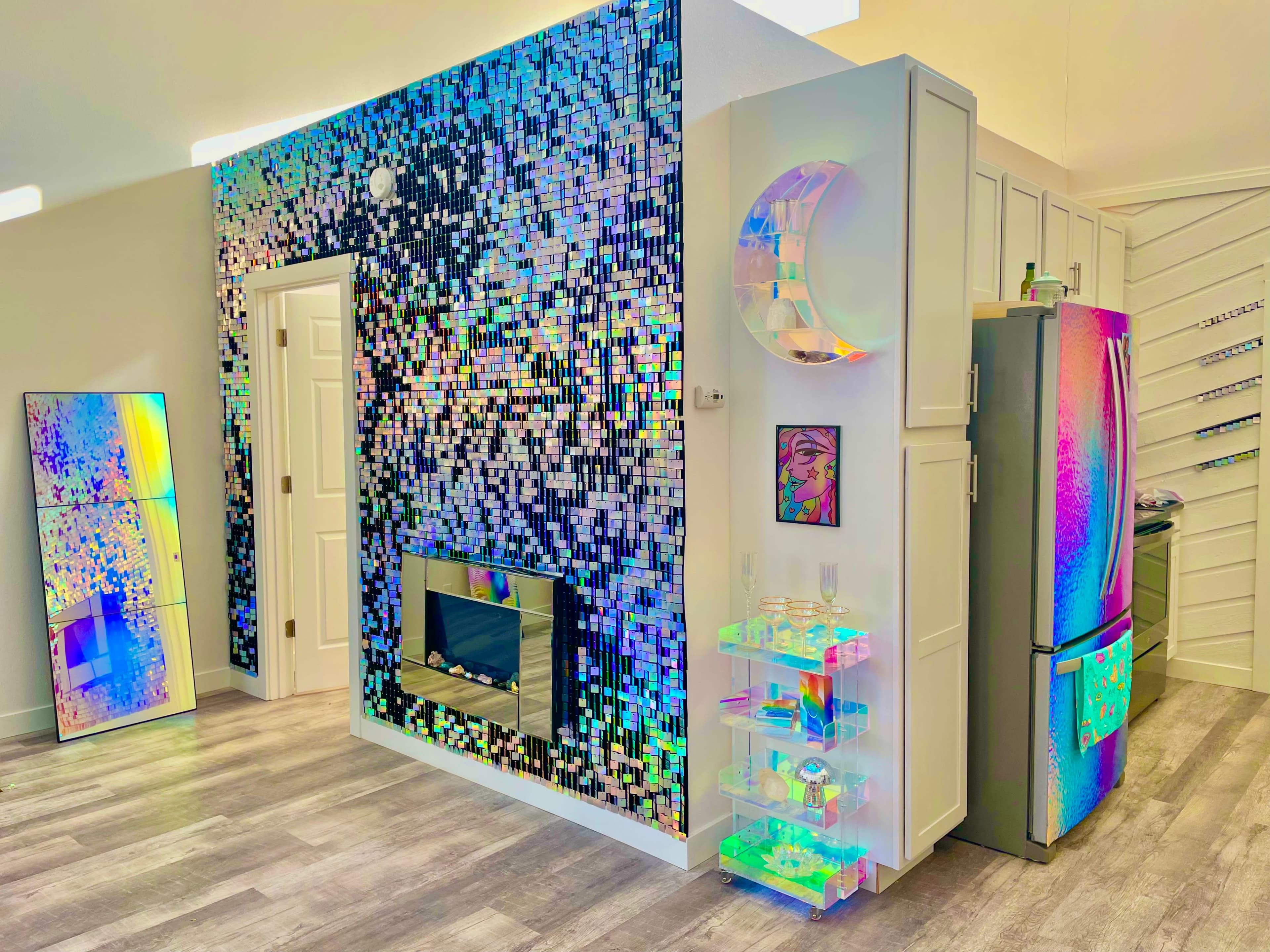 A modern kitchen and living area featuring a reflective mosaic wall, a sleek fireplace, and an iridescent shelving unit next to stainless steel appliances.
