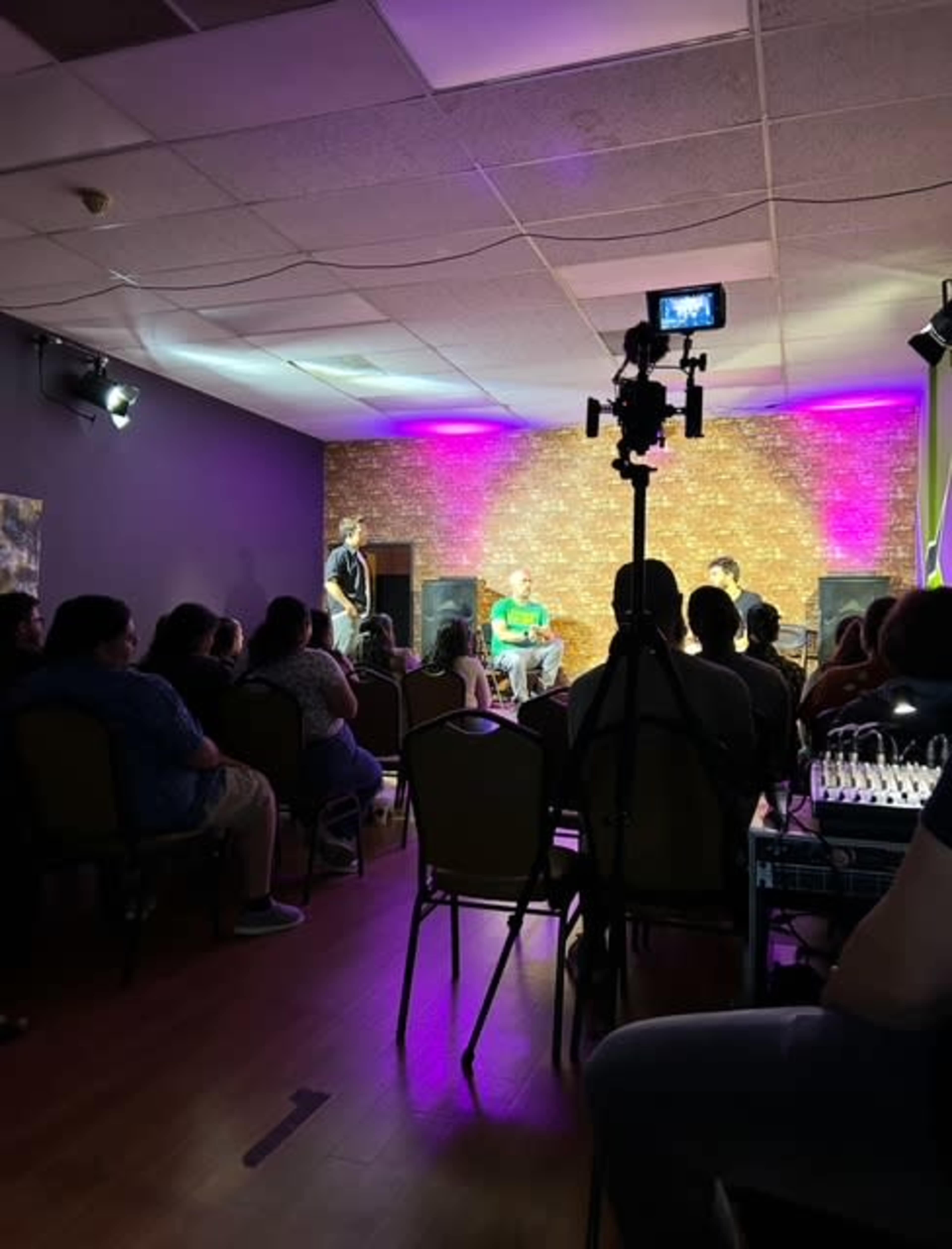 A crowd watches a performance on stage with purple lighting in a room featuring exposed brick walls.