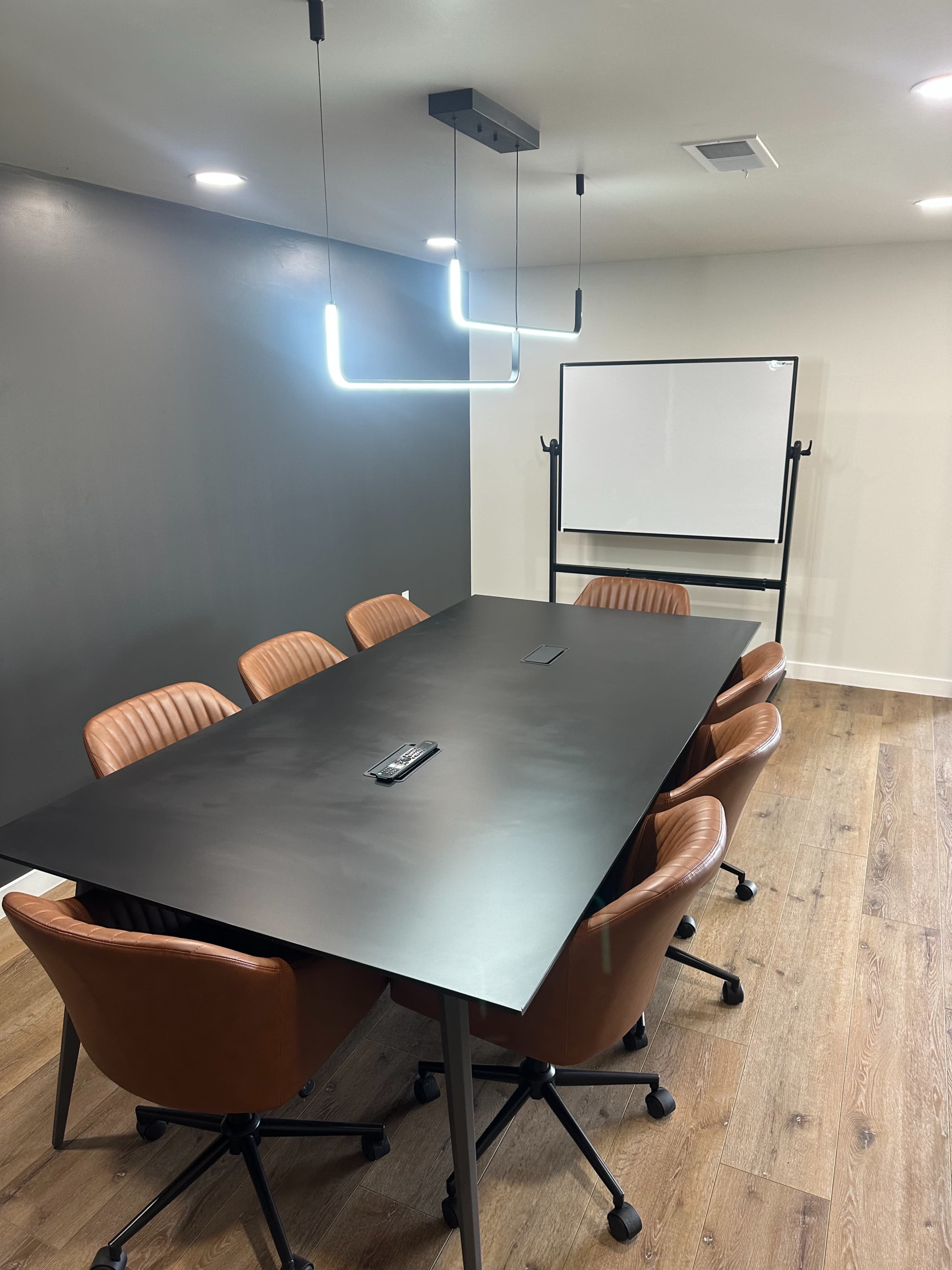 A modern conference room features a large black table surrounded by brown leather chairs, with a projector screen and minimalist lighting.