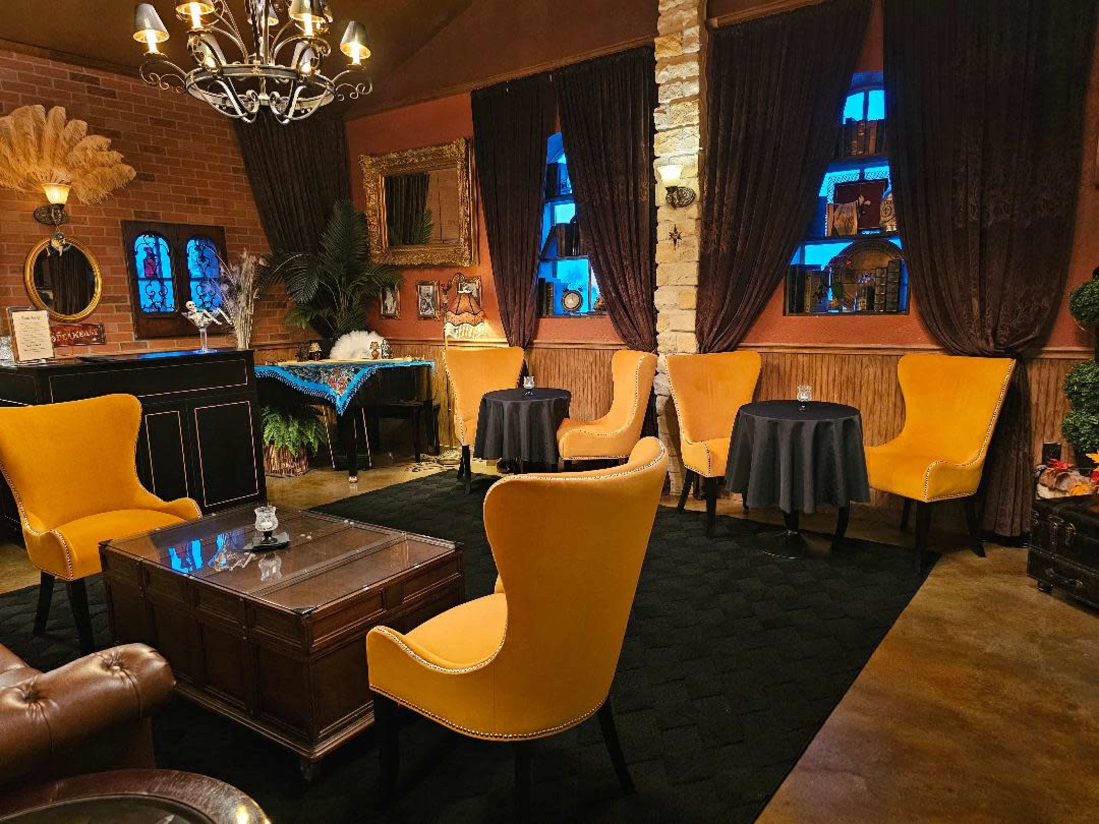 The image shows a cozy lounge area featuring bright yellow chairs, dark wooden tables, and decorative walls, illuminated by a chandelier and soft window light.