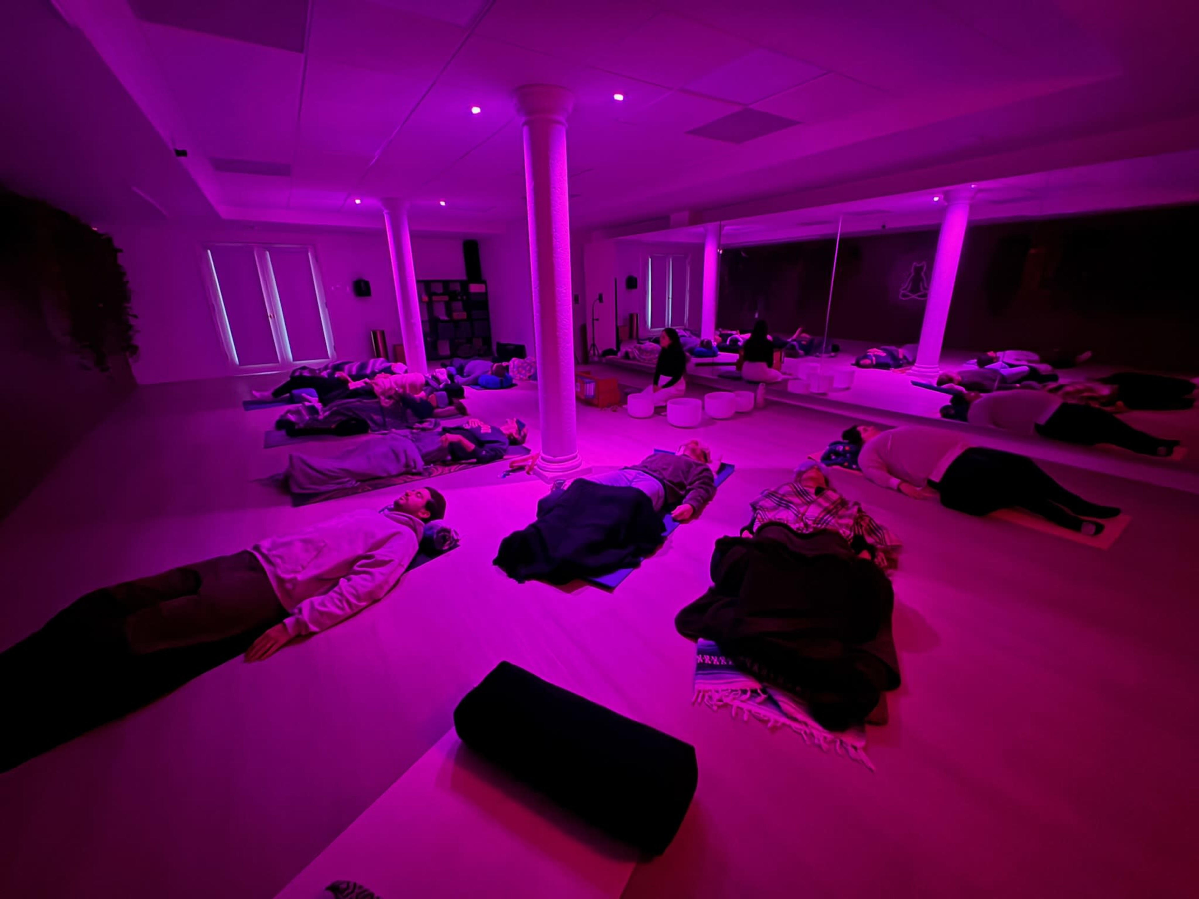 A group of people lie on yoga mats in a softly lit room with pink lighting, while some pillows and cushions are arranged around them.