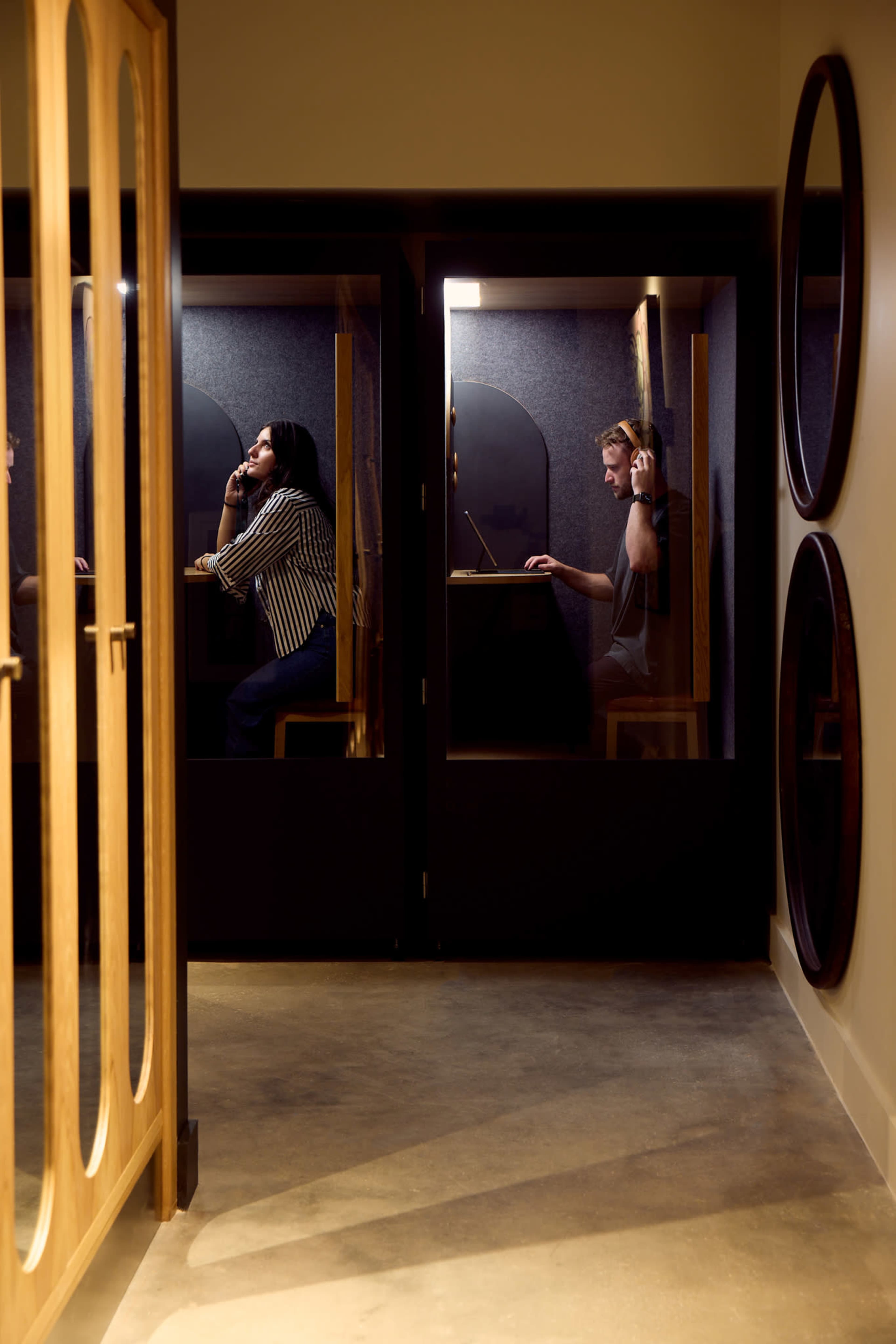 The image shows two people in sound booths, one talking on a phone and the other working on a laptop, with wooden door frames in the foreground.