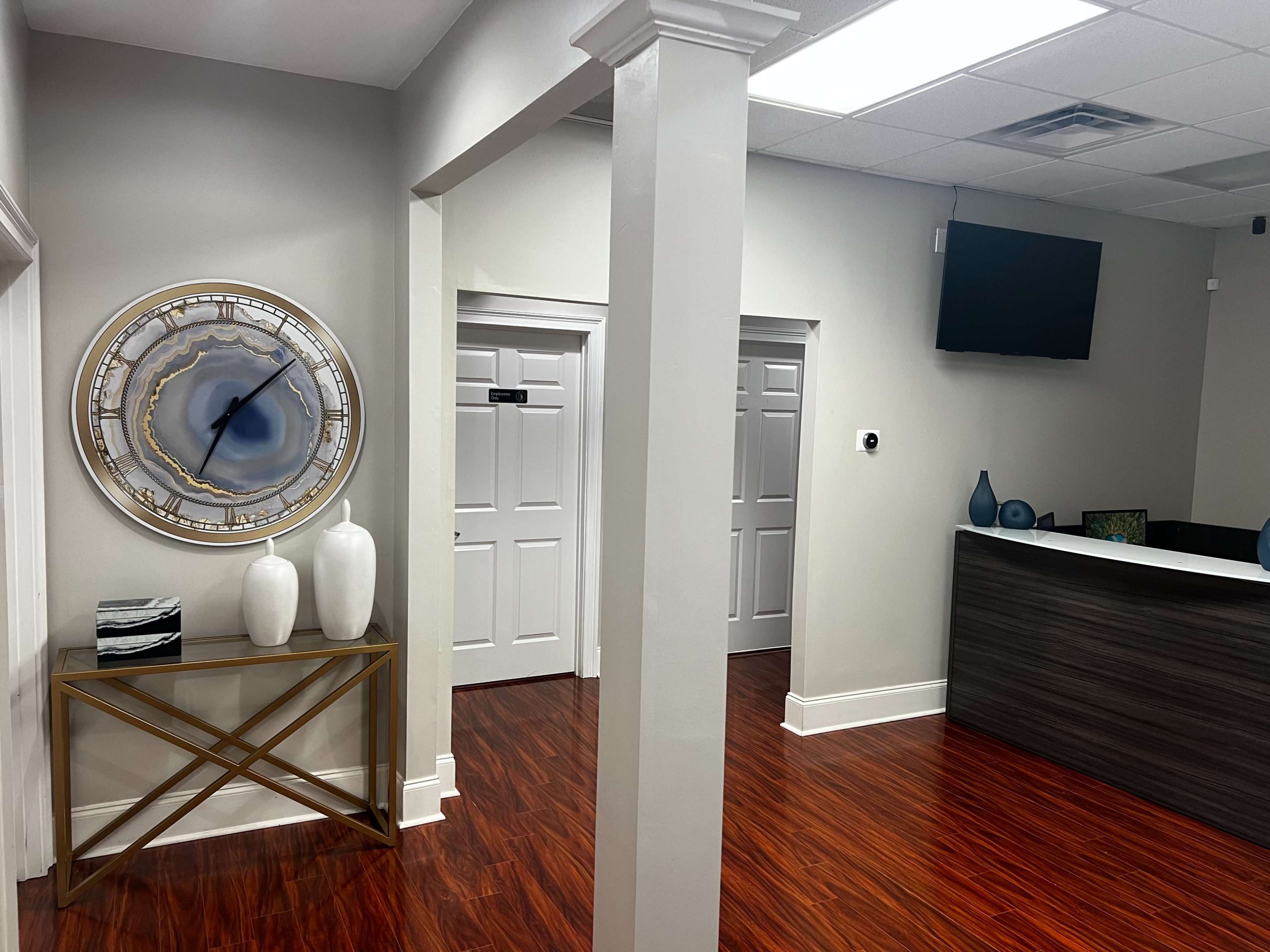 The image shows a modern reception area with a wall clock, two decorative vases on a console table, and a front desk against a backdrop of light-colored walls and wood flooring.