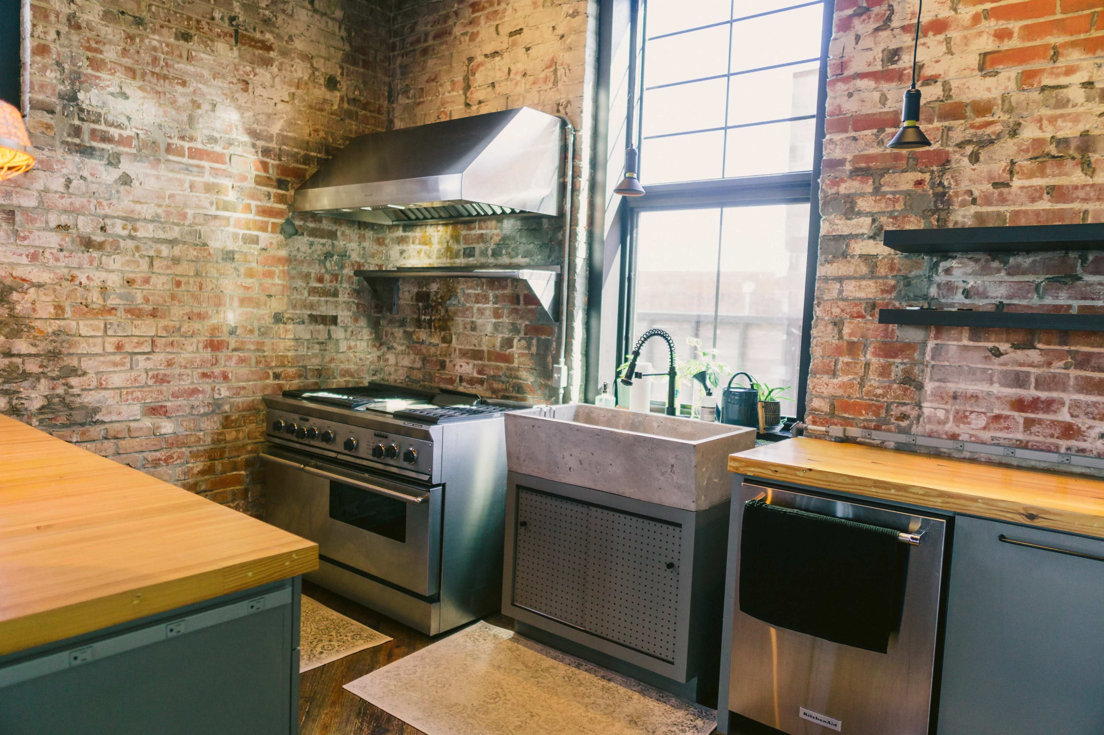 A kitchen with exposed brick walls, a stainless steel range hood above a gas stove, a concrete sink, and wooden countertops.