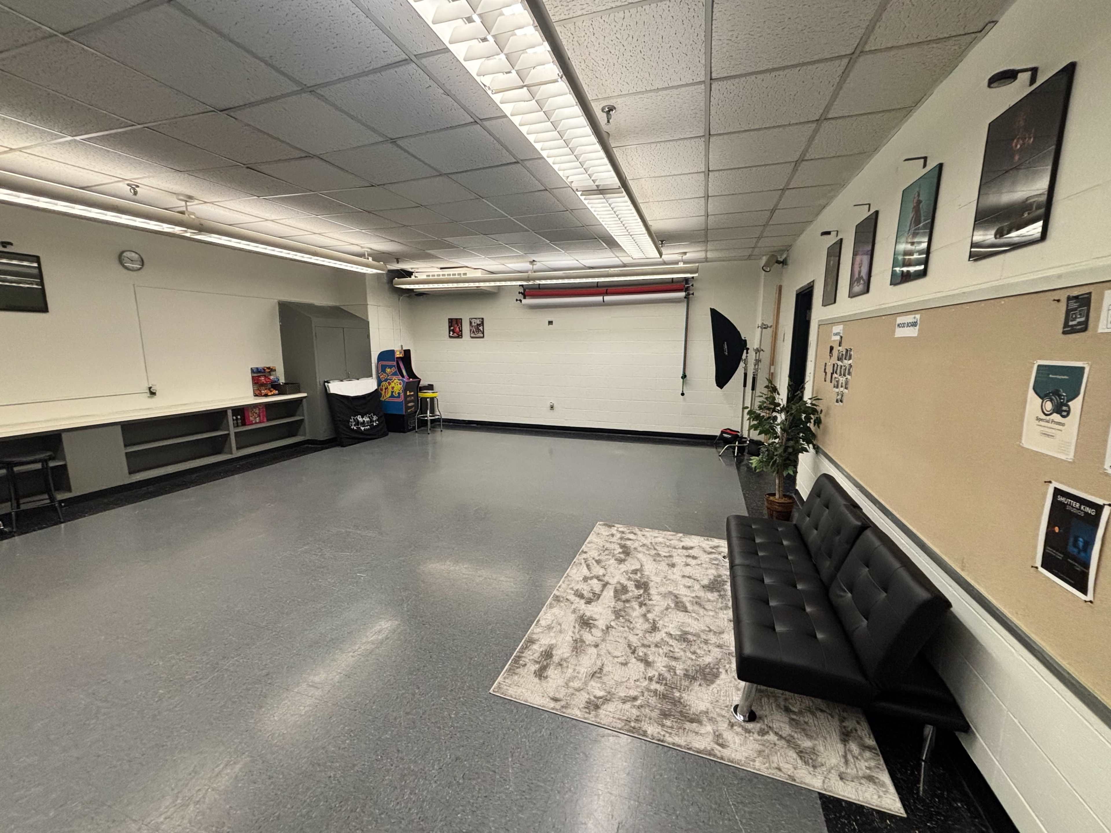The image shows a spacious, empty room with a black sofa, a plant in the corner, a bulletin board on one wall, and various posters hanging on the walls.