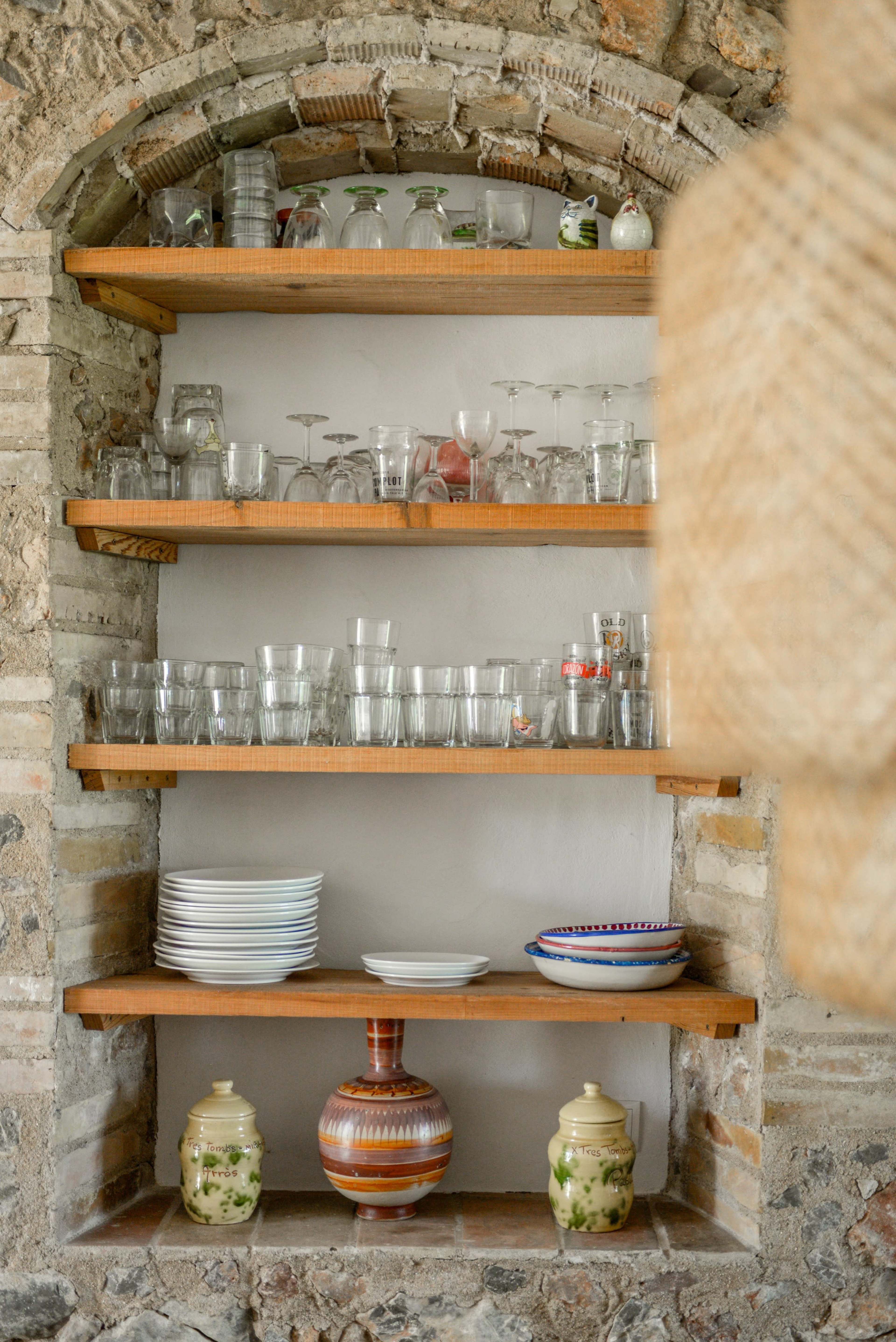 A wooden shelving unit displays various glassware and dishes in a stone wall alcove.