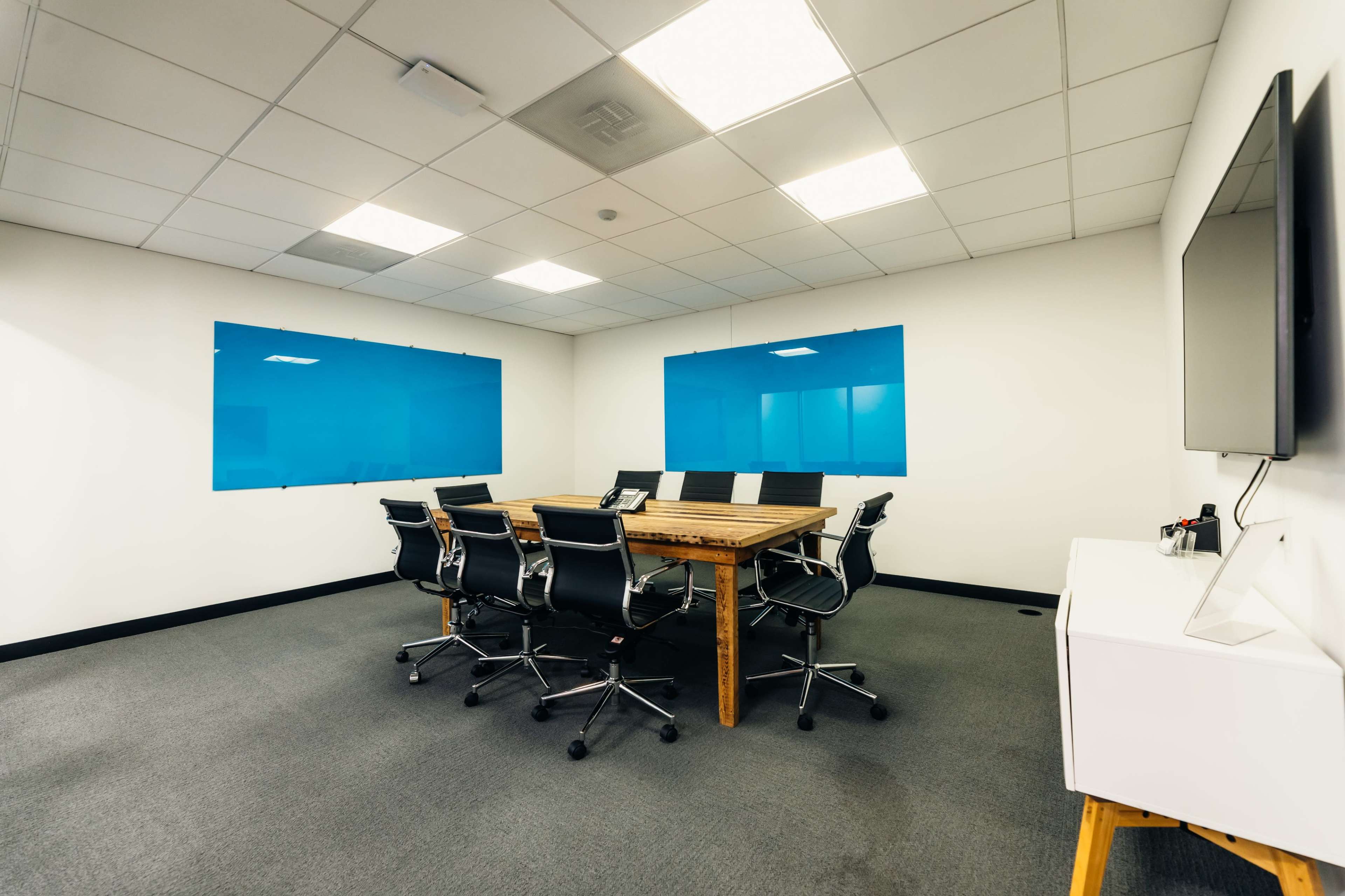 A conference room features a large wooden table surrounded by black office chairs, with blue panels on the walls and a white storage unit.