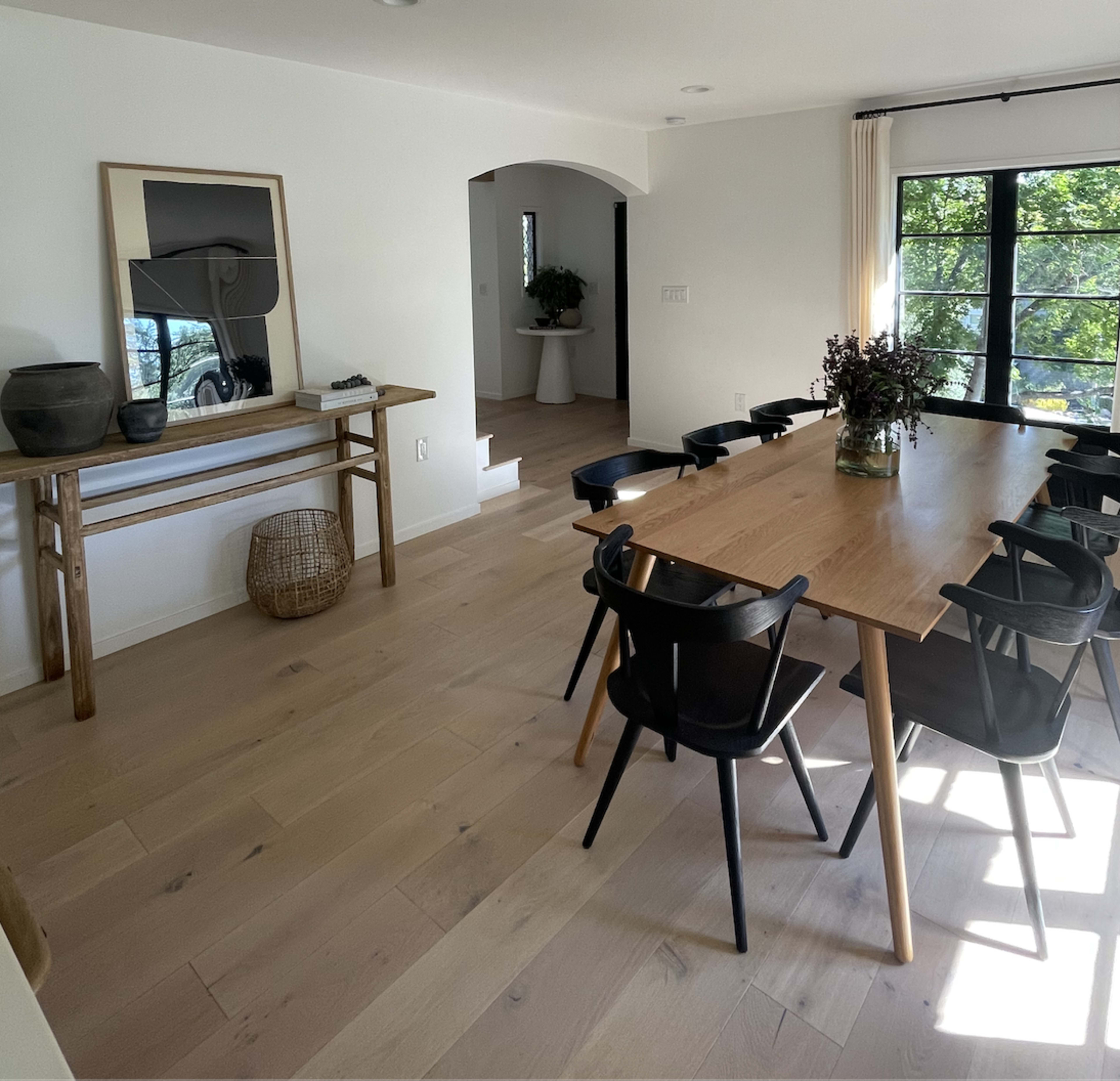 The image shows a modern dining area featuring a wooden table surrounded by black chairs, with a side console table and a vase of flowers on display.
