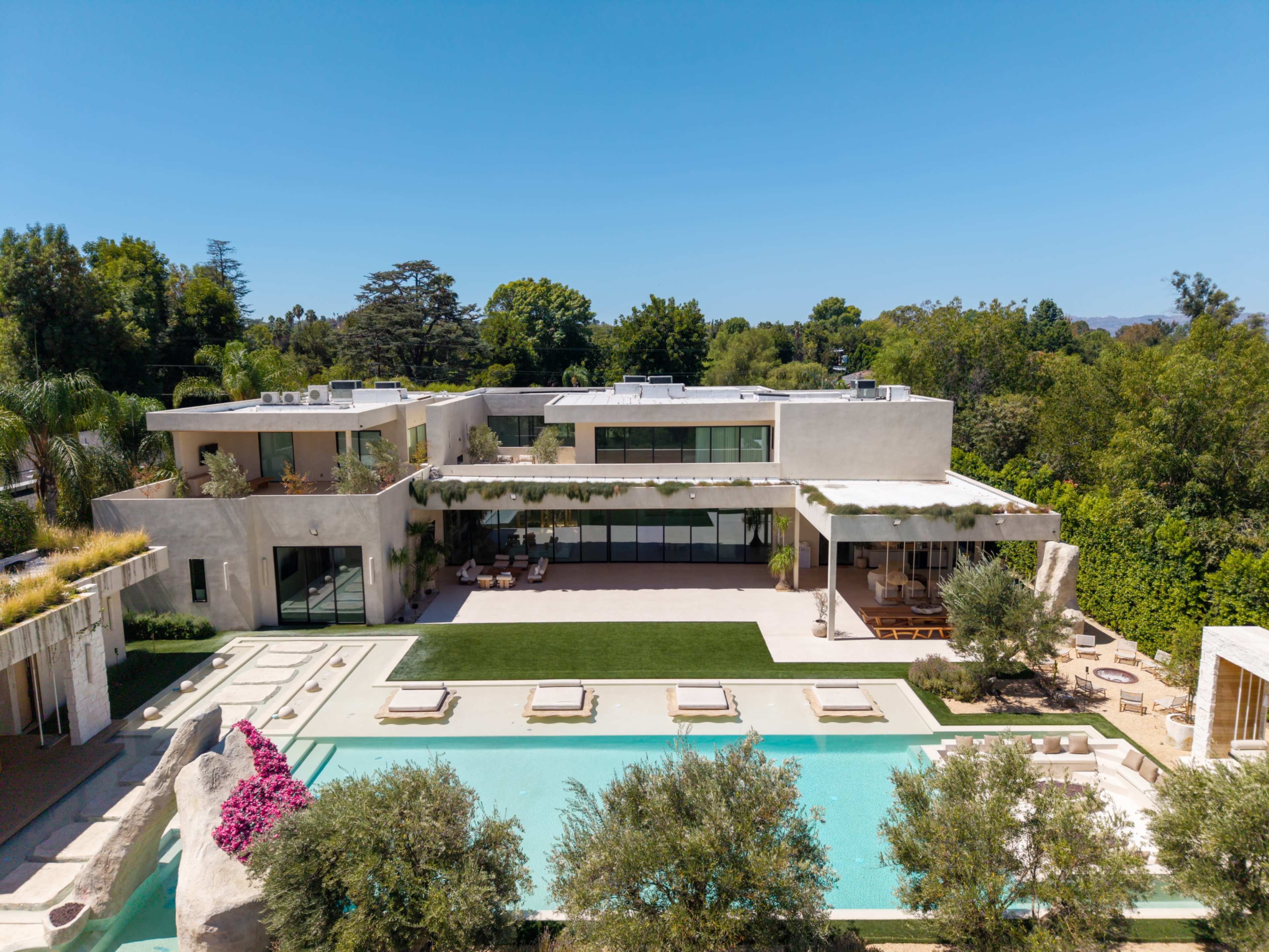 The image shows a modern, multi-level house with large windows, a swimming pool, and landscaped gardens surrounded by trees.