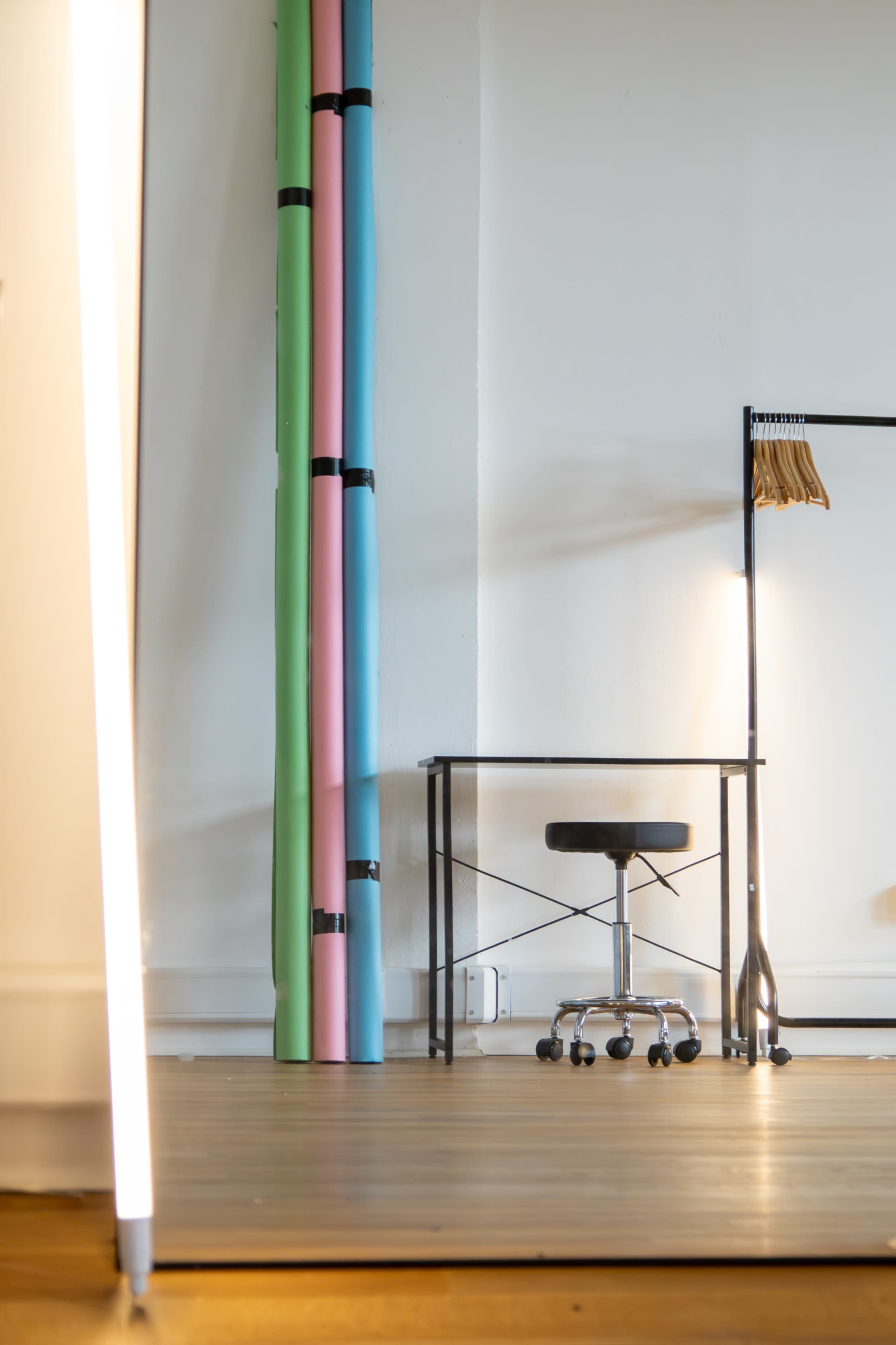 The image shows a minimalist studio space featuring a black rolling stool, a metal table, and colorful paper rolls leaning against the wall.