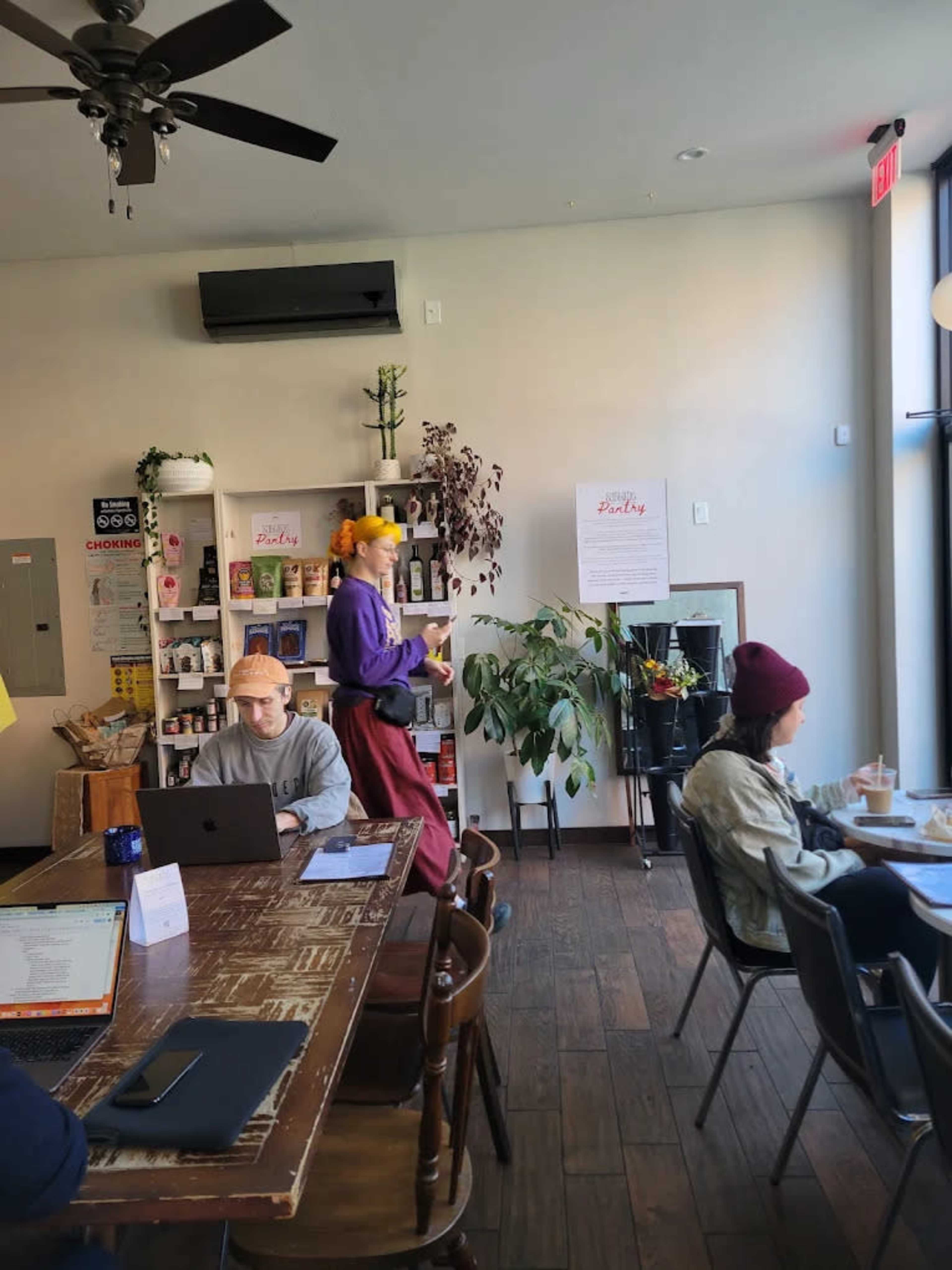 A coffee shop scene features a person in bright attire walking by a table where two customers are seated, with shelves of products in the background.