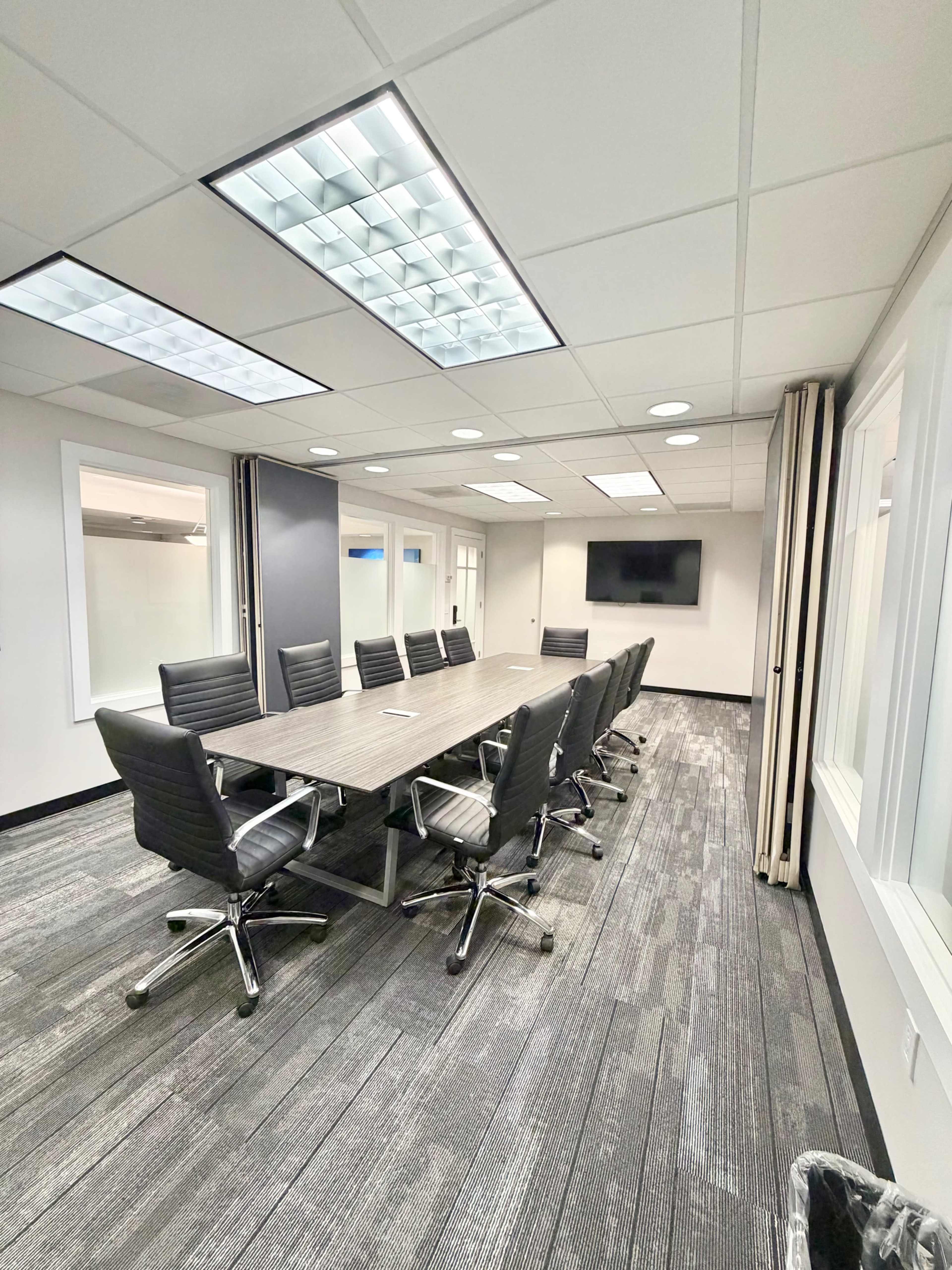 The image shows a modern conference room with a large rectangular table surrounded by ergonomic chairs, featuring a wall-mounted screen and large windows.