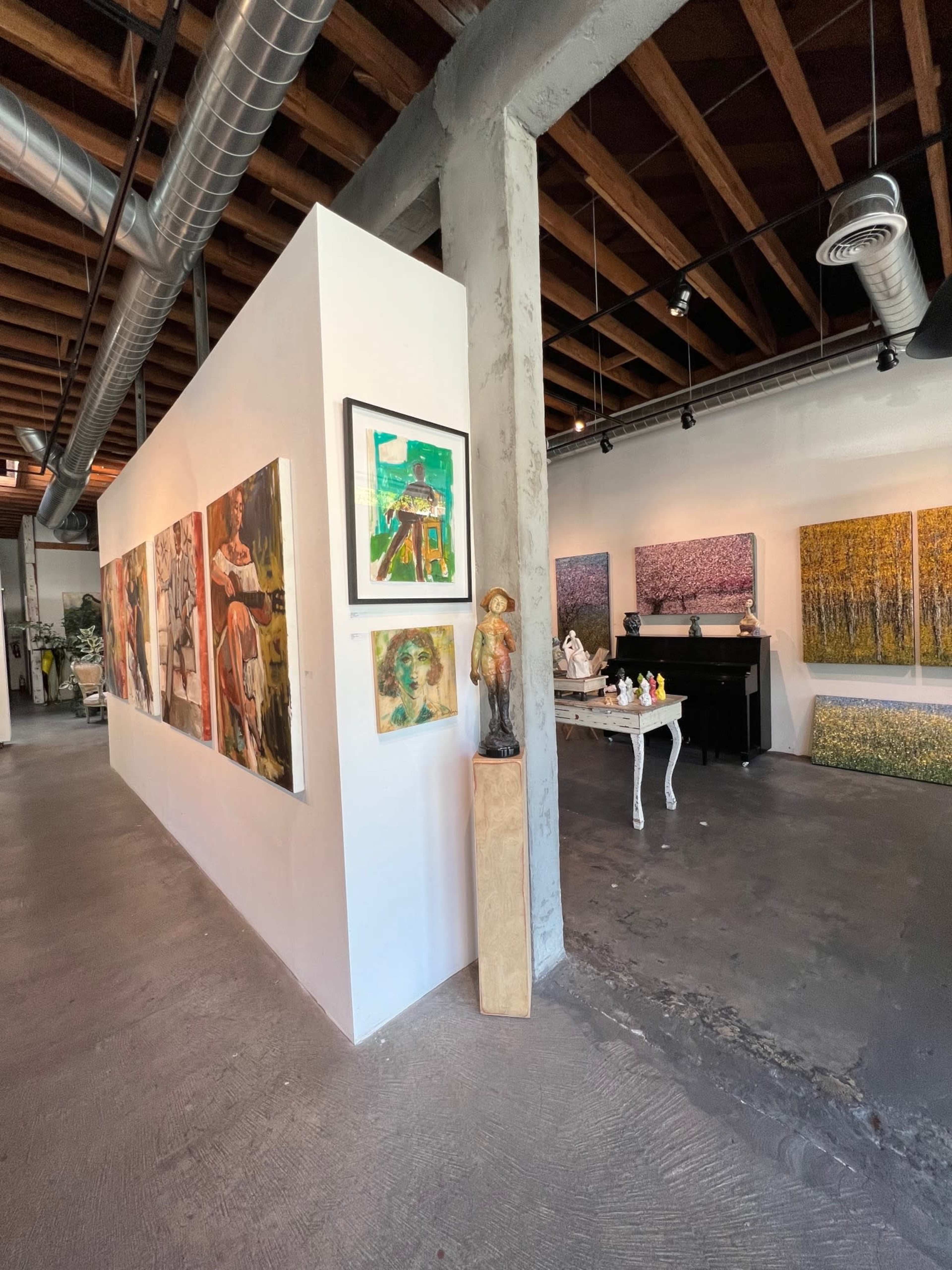 An art gallery interior featuring a series of framed paintings on a white wall and various artworks displayed on tables and stands.