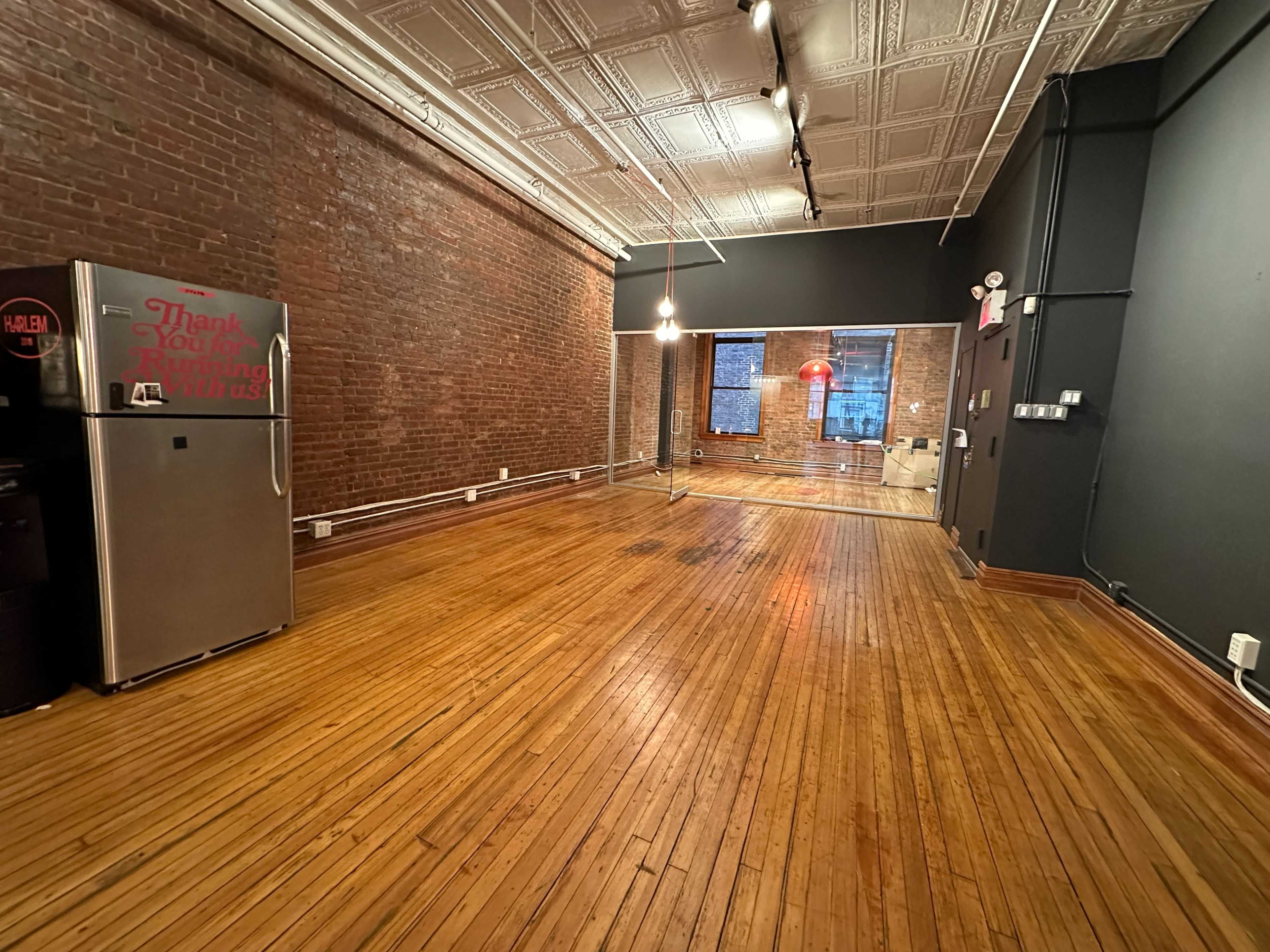 An empty room features exposed brick walls, polished wooden floors, and a large window displaying a red neon light.