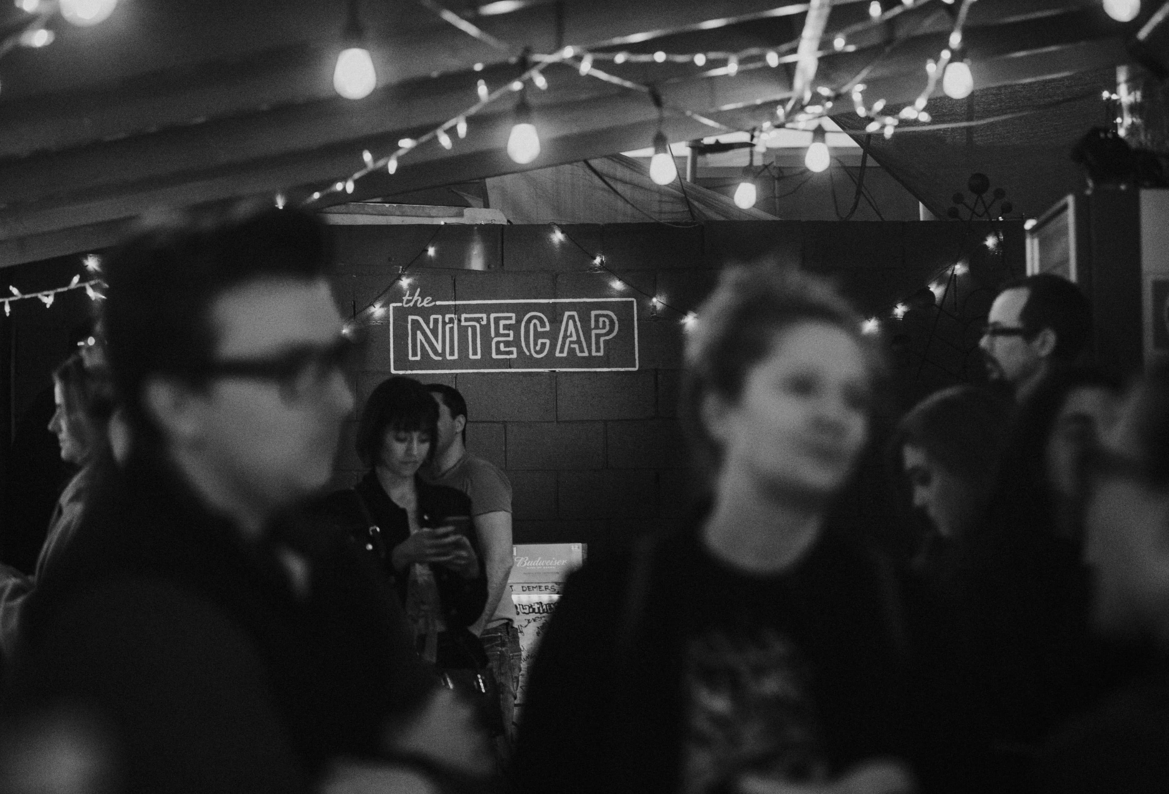 A dimly lit venue named "the Nitecap" is filled with people chatting and standing under string lights.