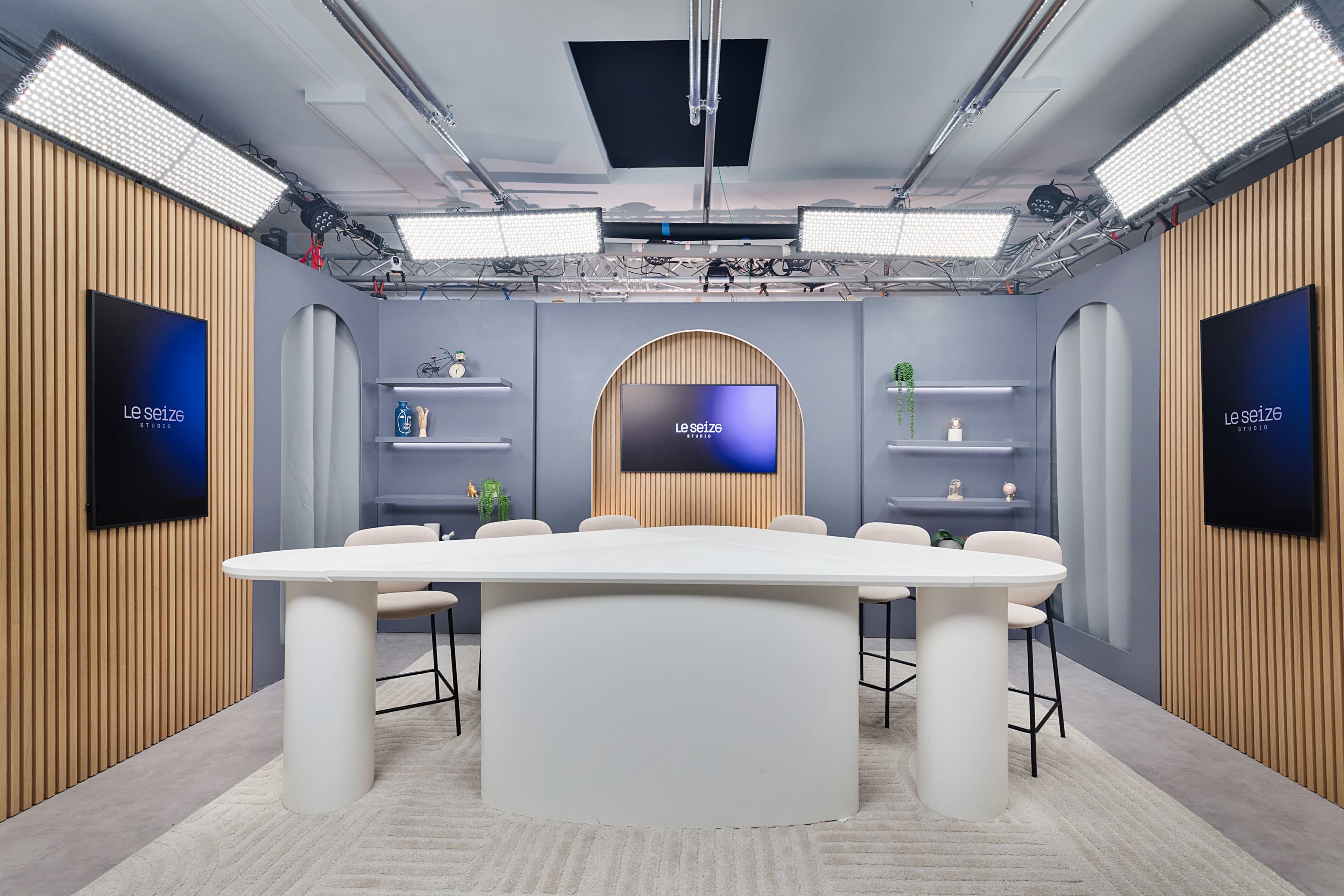 The image shows a modern studio set with a large white table surrounded by barstools, featuring multiple screens and shelves with decorative items.