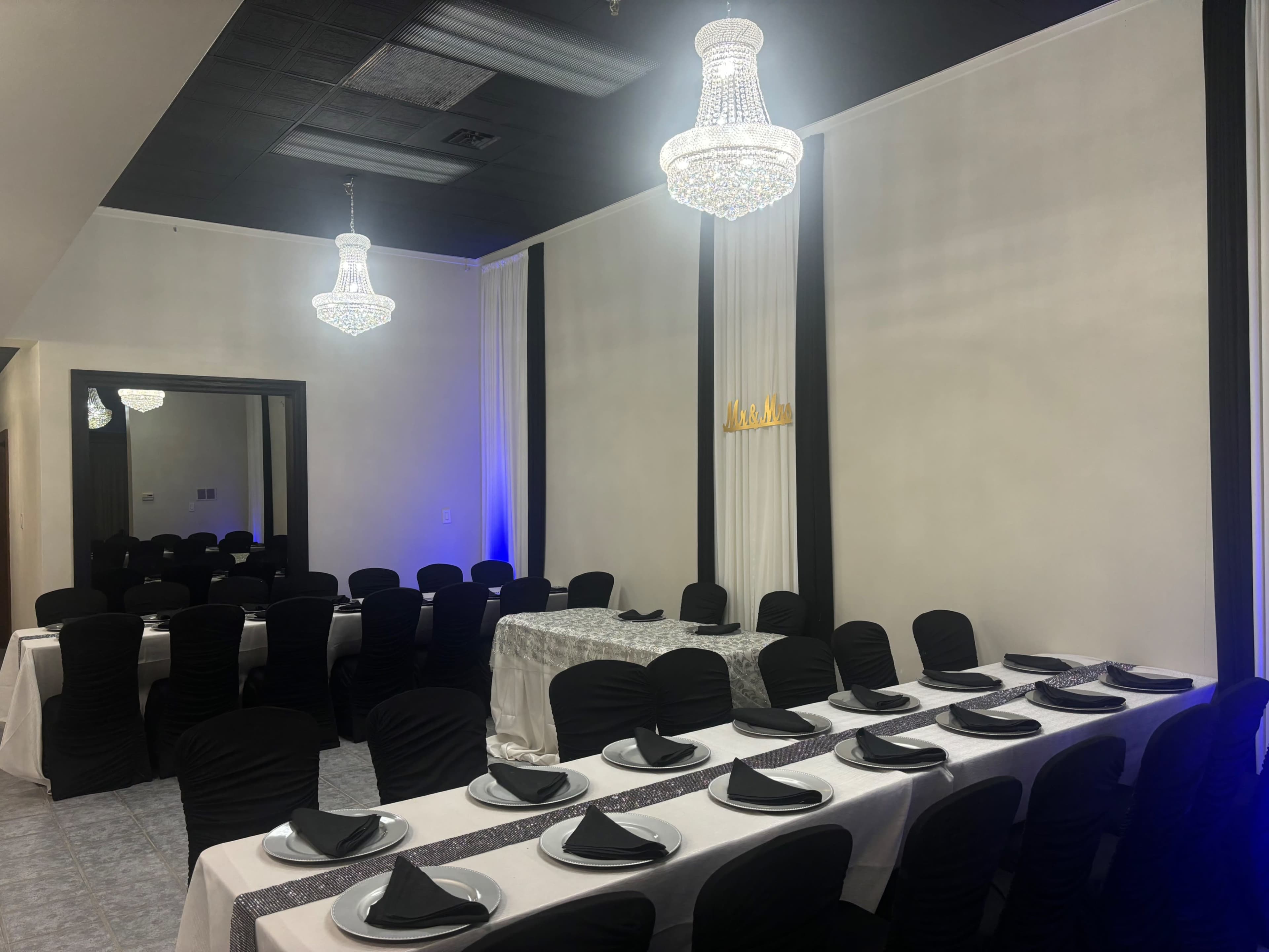 The image shows a room set up for a formal dining event, featuring black chairs with white tables arranged in rows, adorned with black napkins and chandeliers overhead.