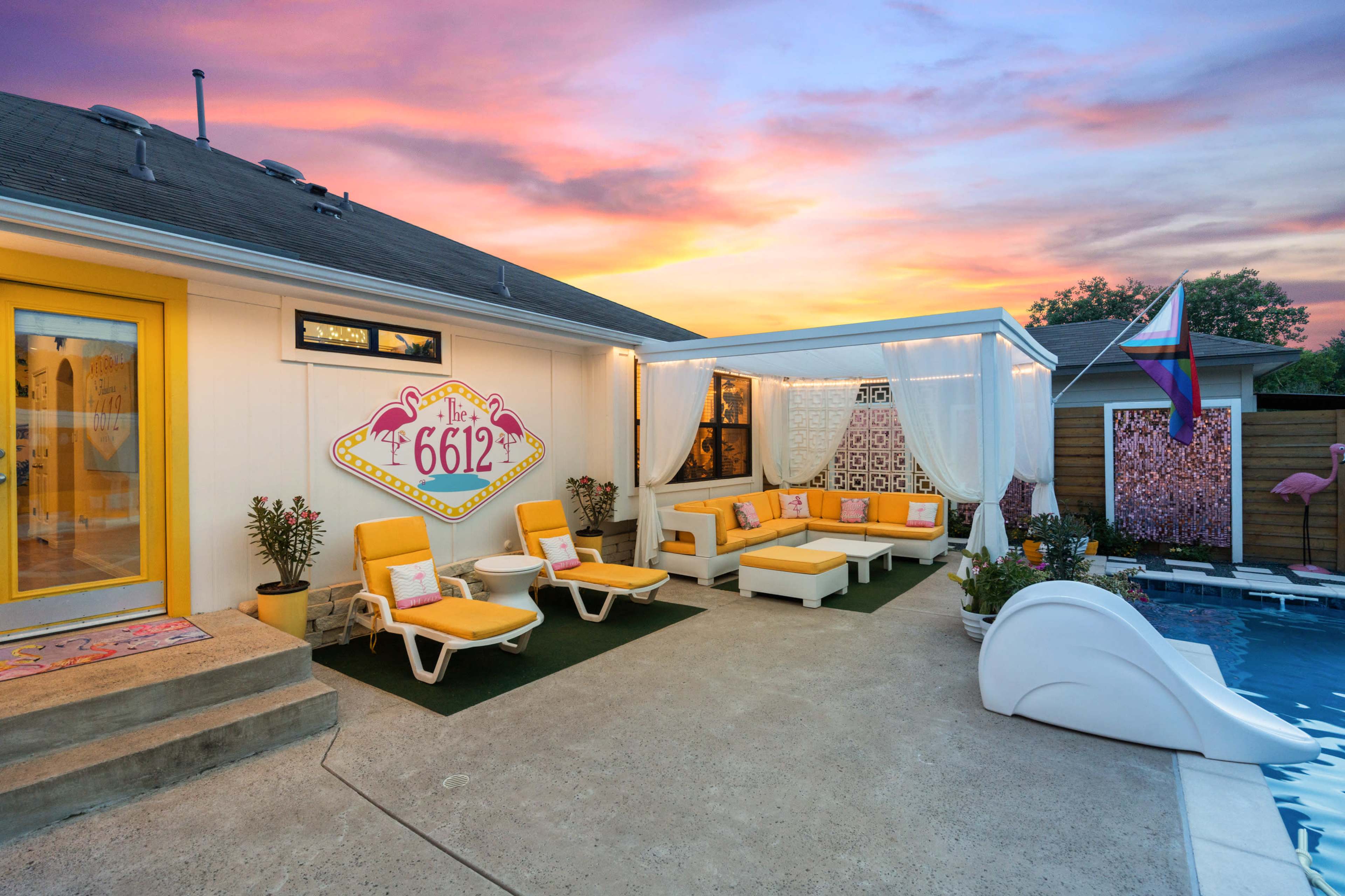 The image shows a vibrant outdoor seating area with yellow lounge chairs, a white sectional sofa, and a pool against a colorful sunset sky.