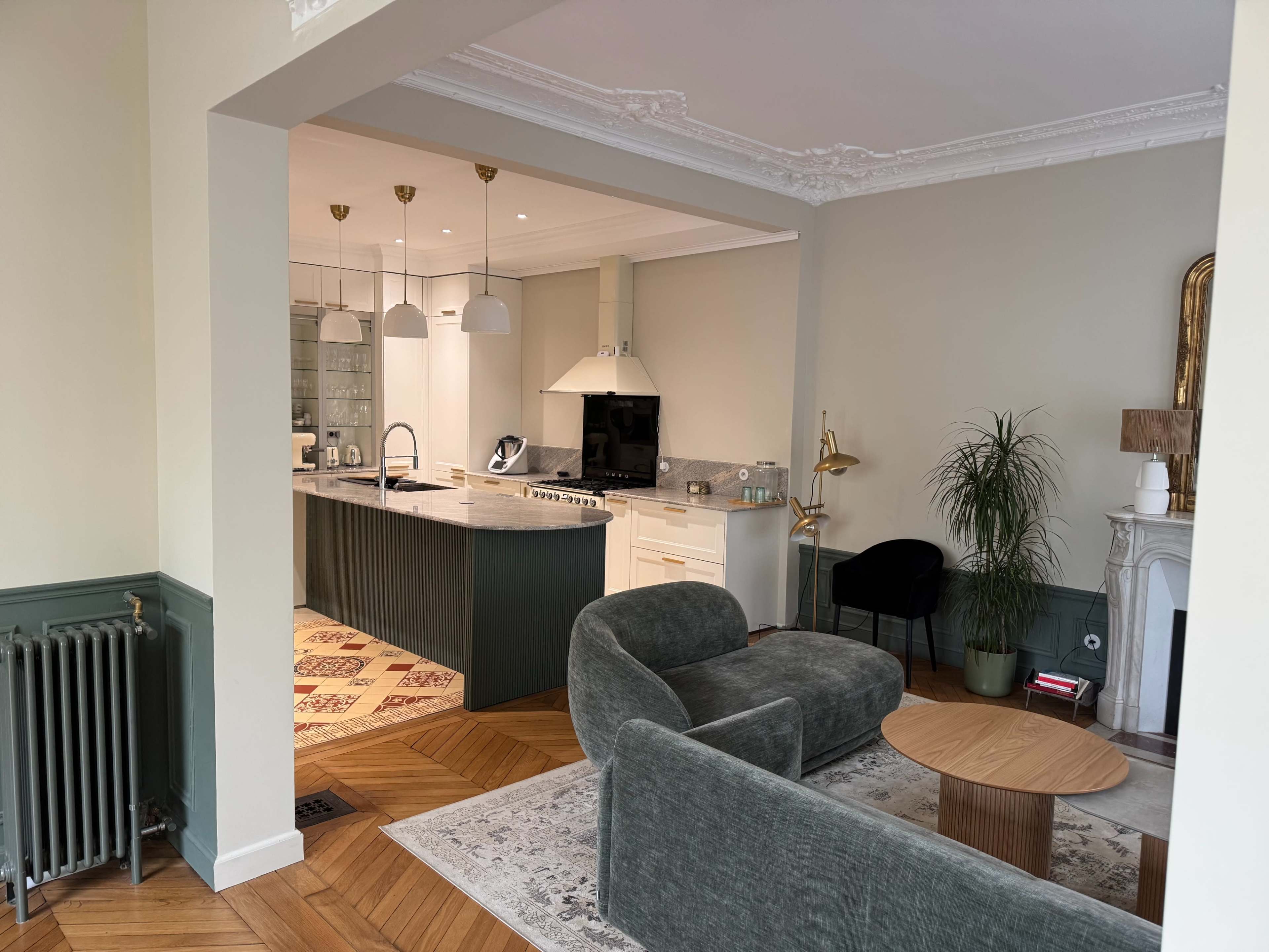 The image shows a modern living room with a green sofa, a round wooden coffee table, and a view of a kitchen area featuring white cabinets and a marble countertop.