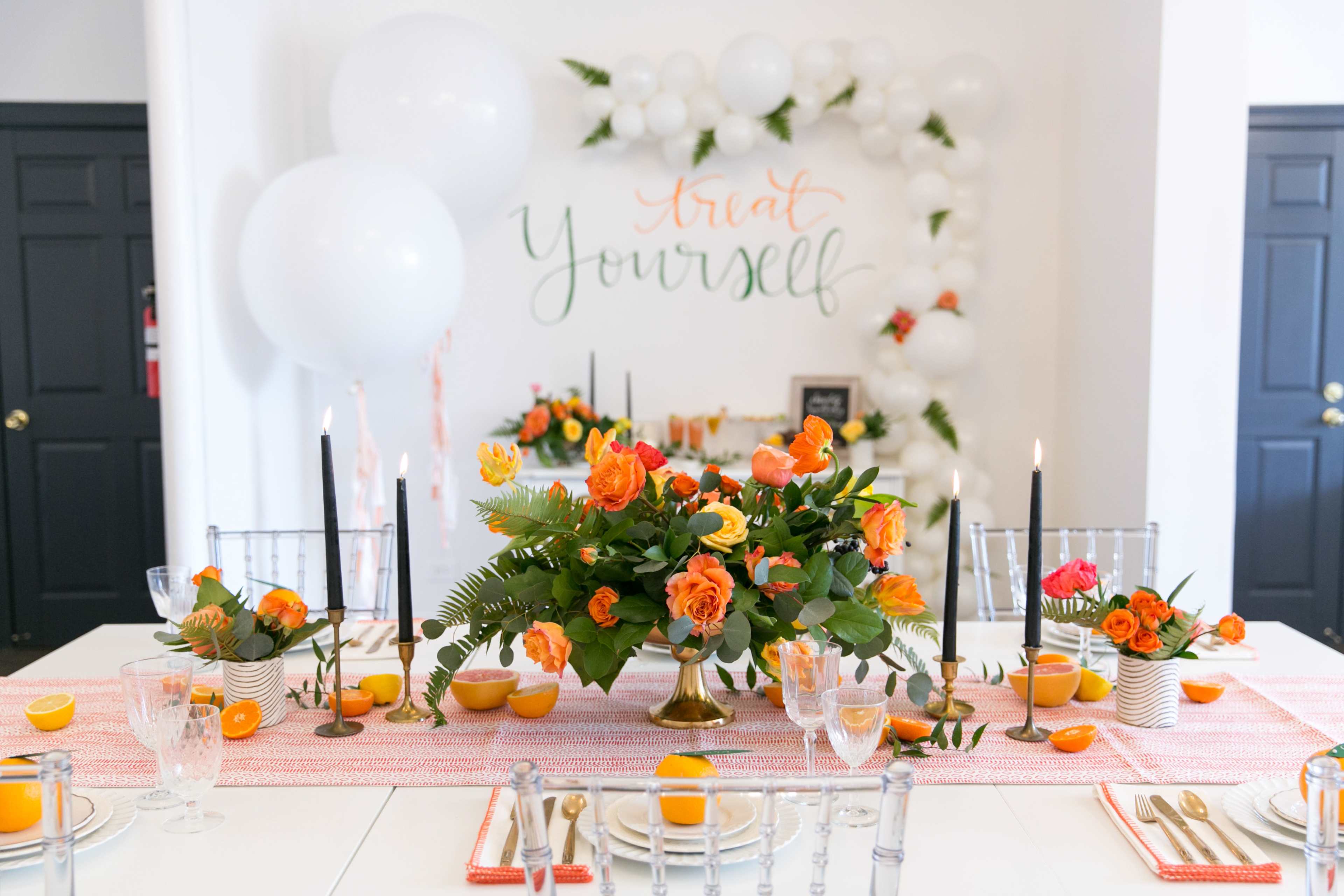 A decorated dining table features an orange floral centerpiece, black candlesticks, and vibrant place settings, set against a backdrop with the words "Treat Yourself."