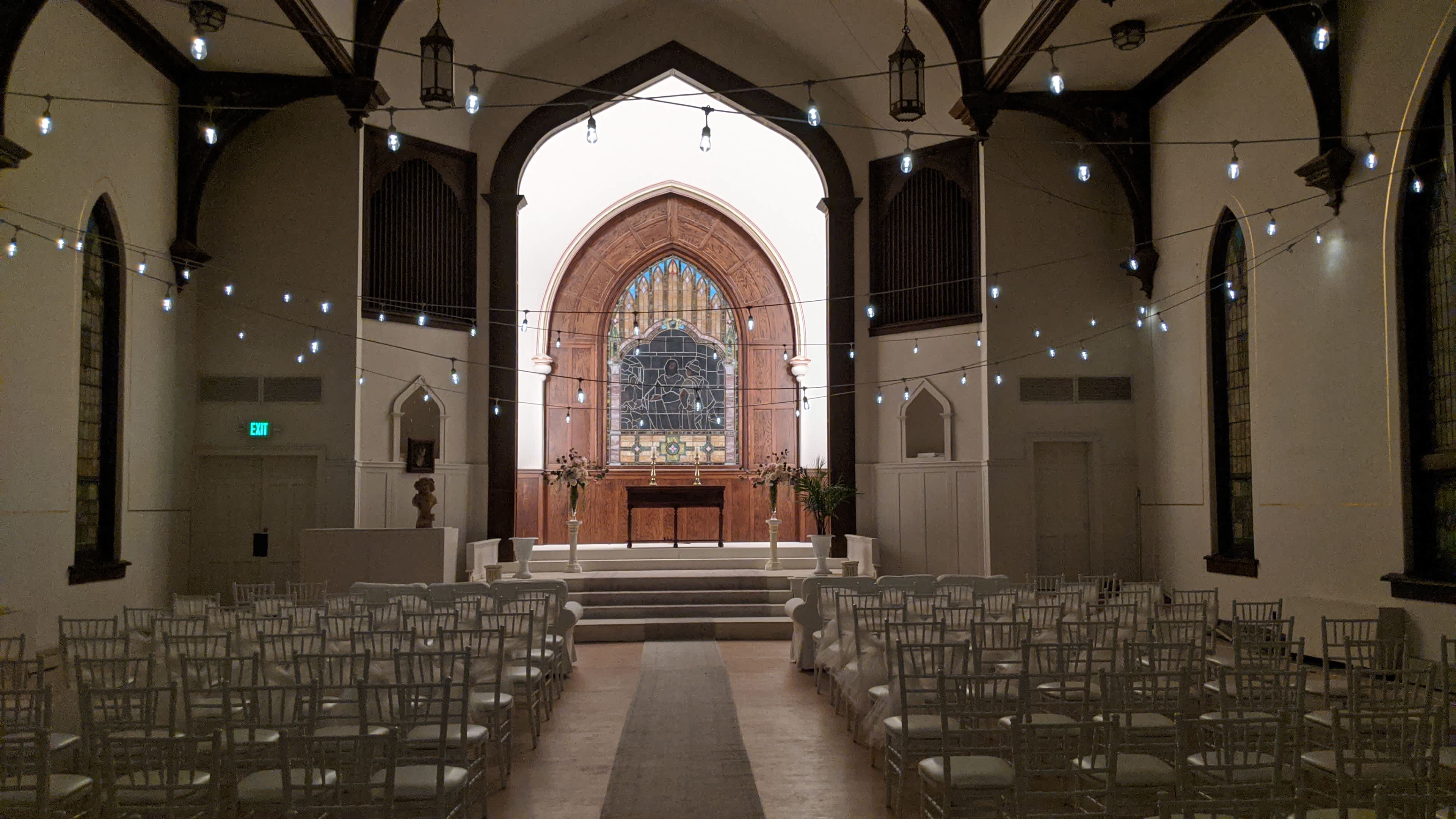 An interior of a church or event space with rows of chairs facing a wooden altar, illuminated by strings of lights and featuring a large stained glass window at the back.