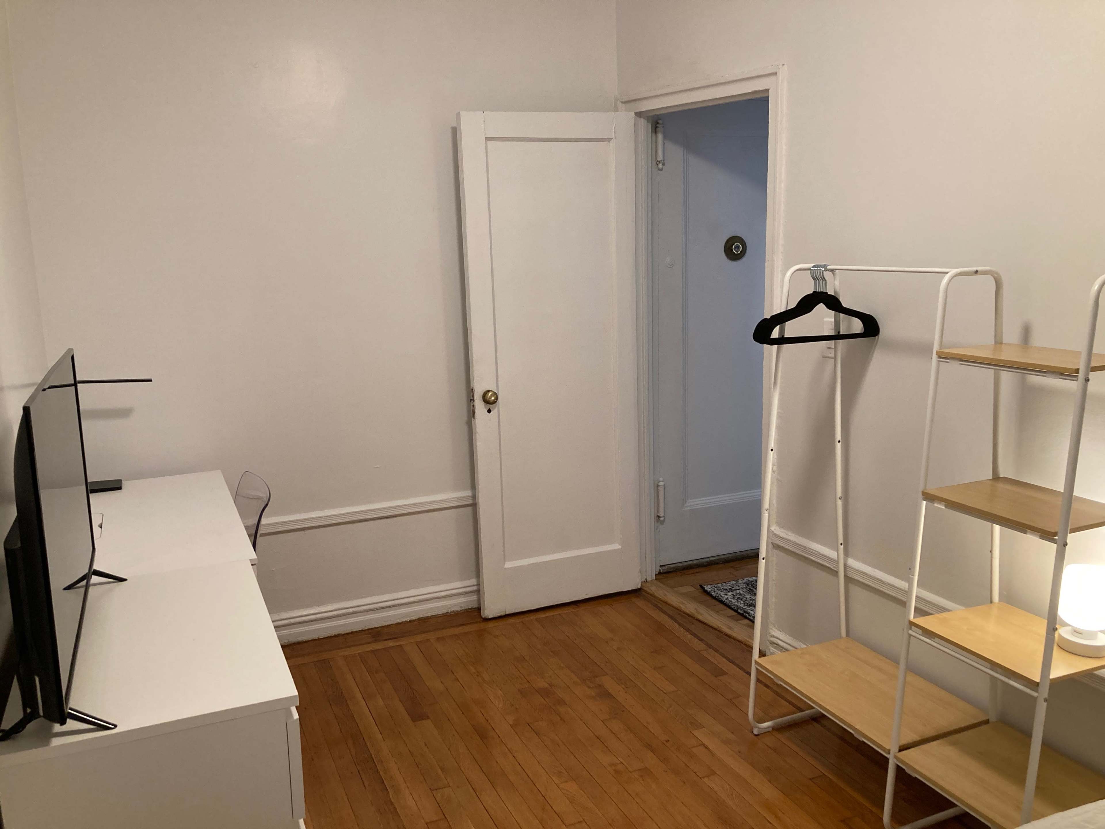A small room with a white wall, a television stand, a coat hanger, and a wooden shelf, along with a door leading to a hallway.