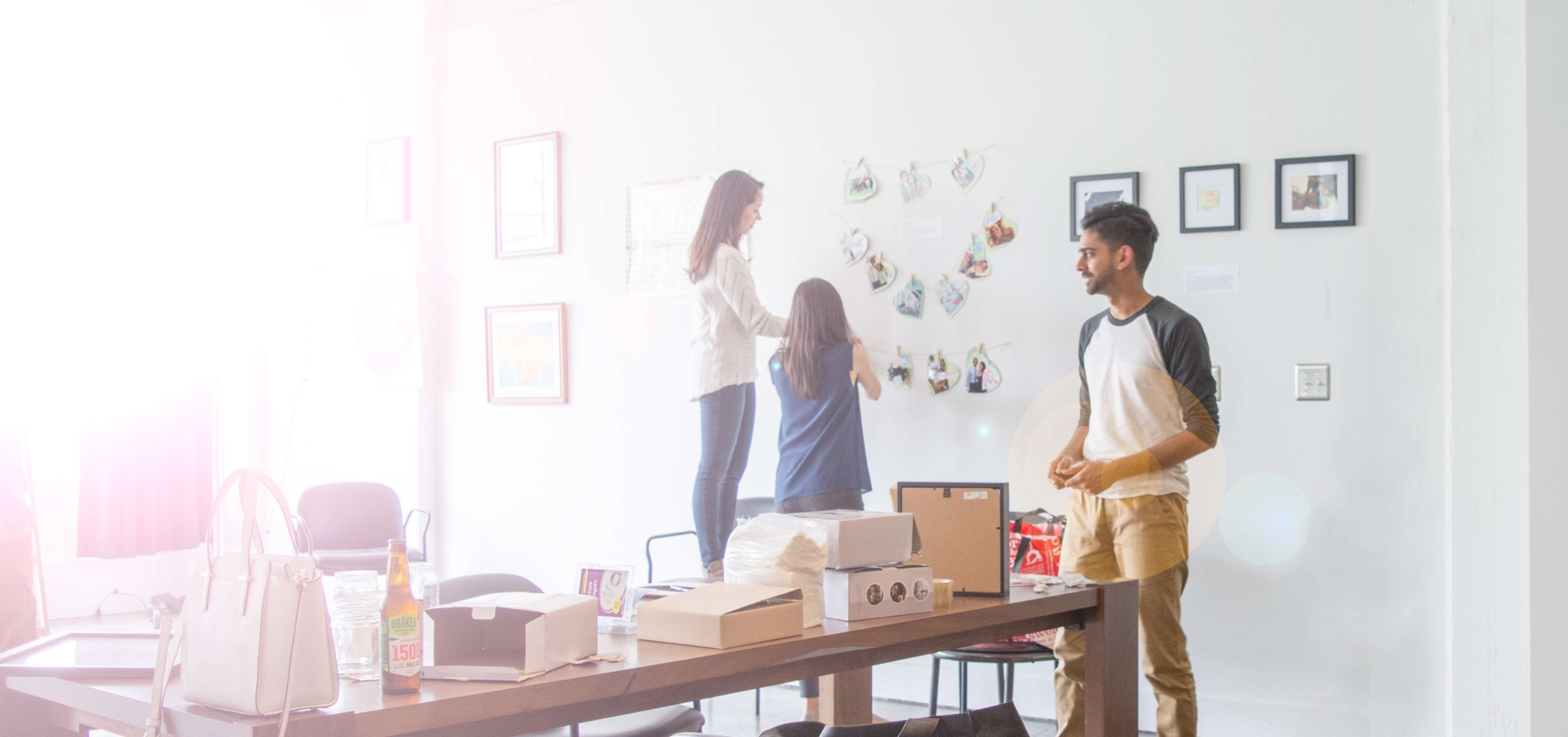 Two people are hanging photos on a wall while a third individual stands by a table in a bright, open room.