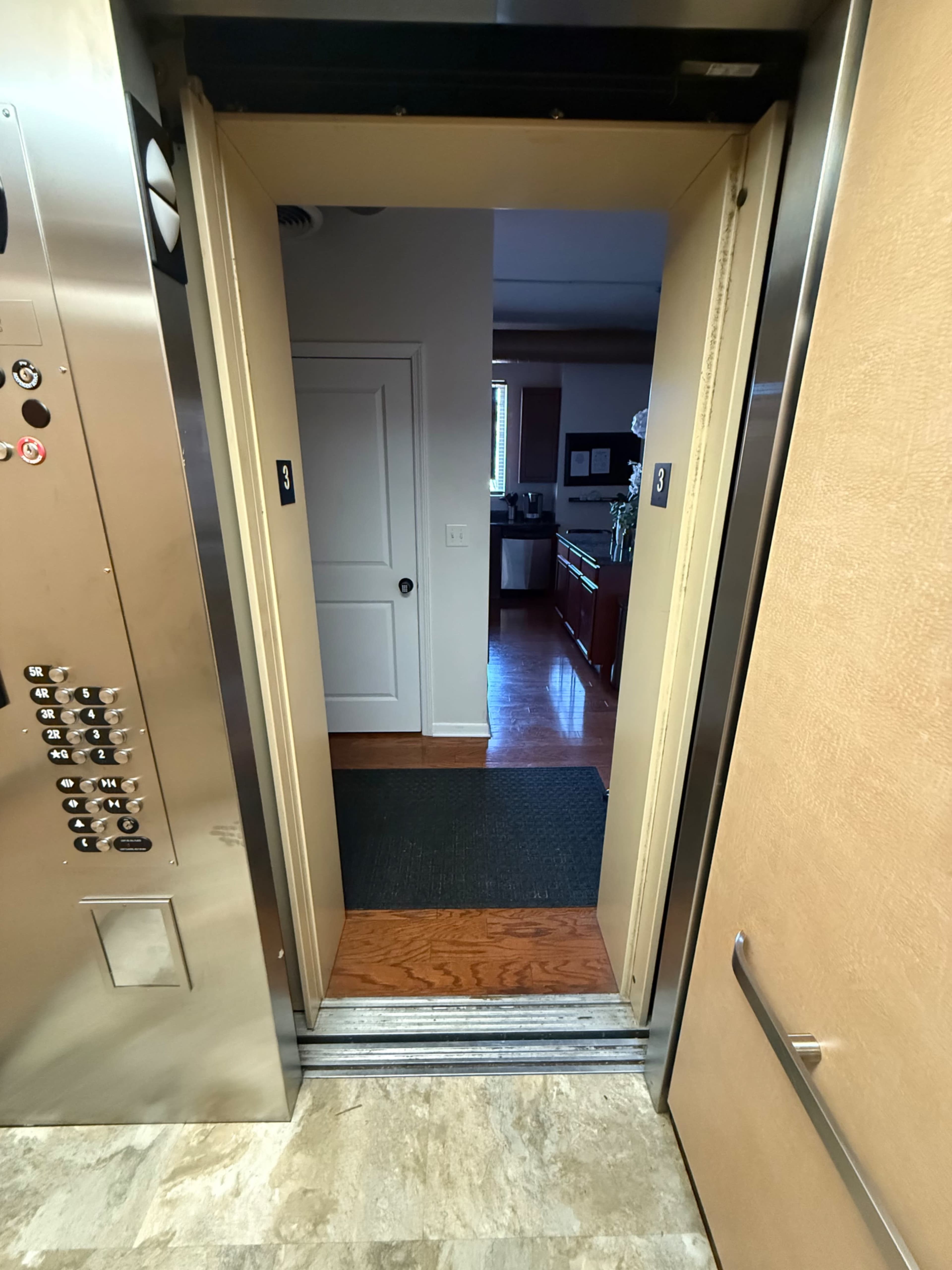 The image shows the open doors of an elevator revealing a hallway with a door at the end and wooden flooring.