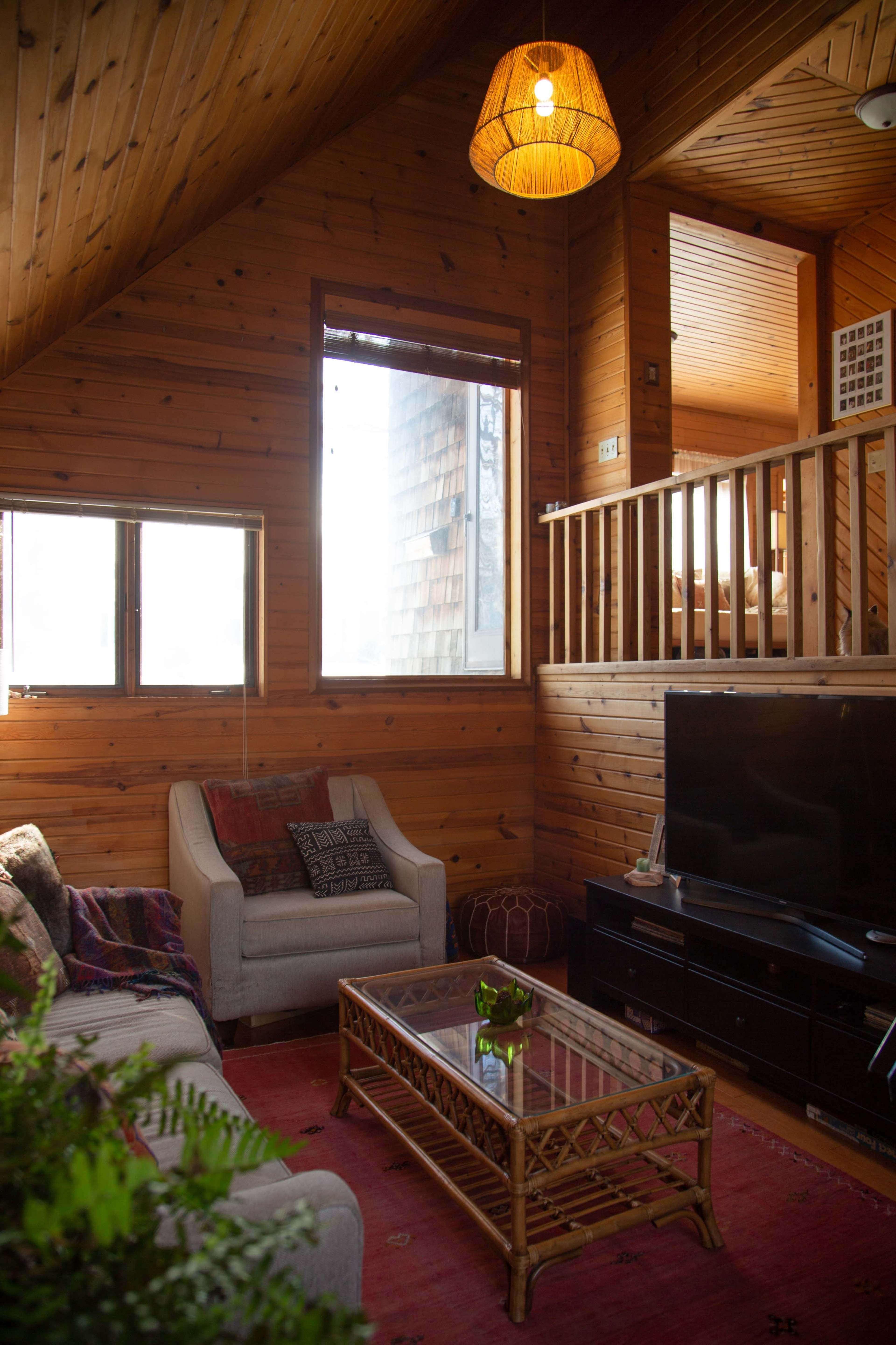 A wooden interior living space with a sofa, a coffee table, and a television, featuring a large window that lets in natural light.