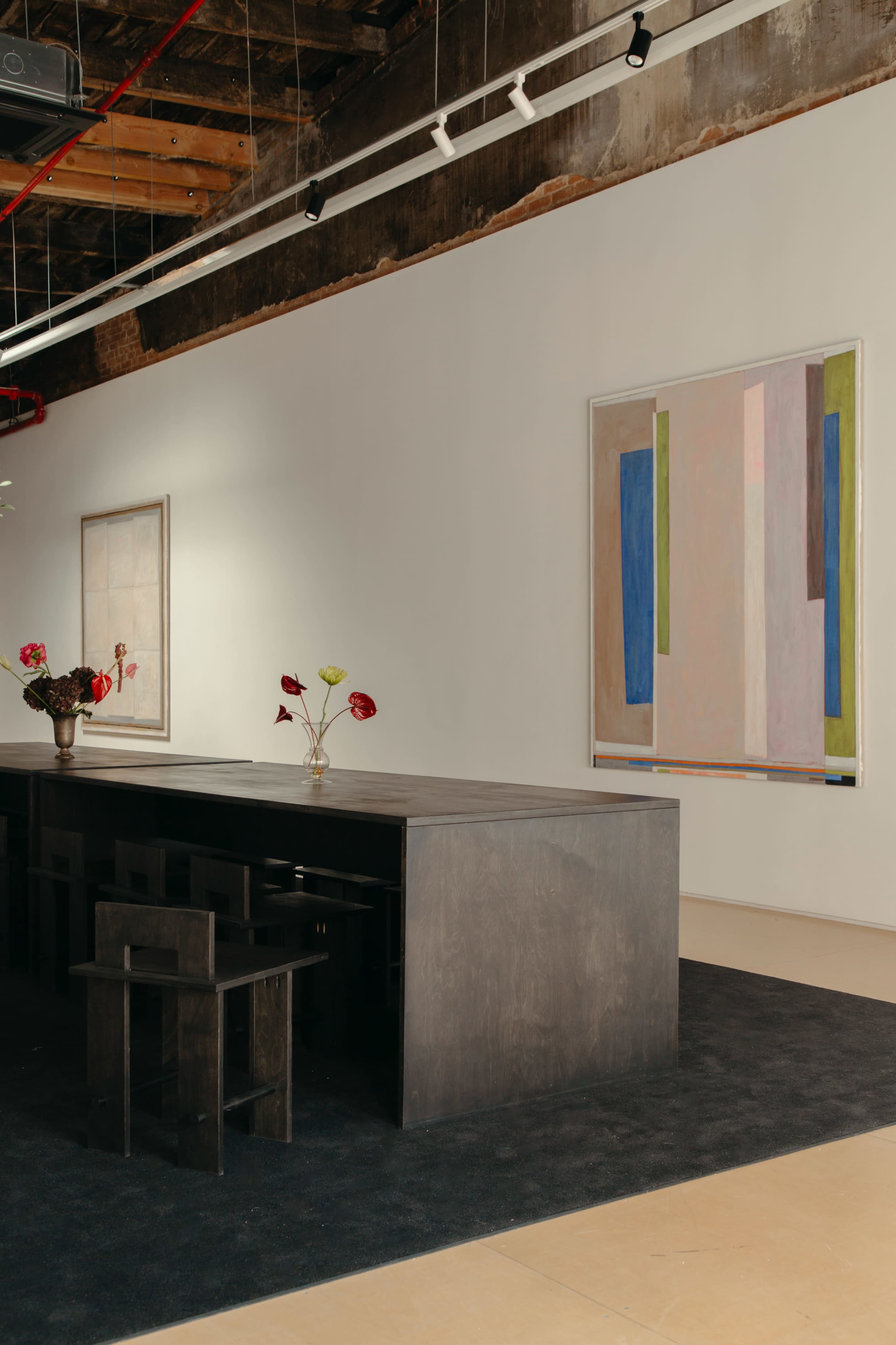 The image shows a modern art gallery interior with a long black table, black chairs, and abstract paintings displayed on the white walls.