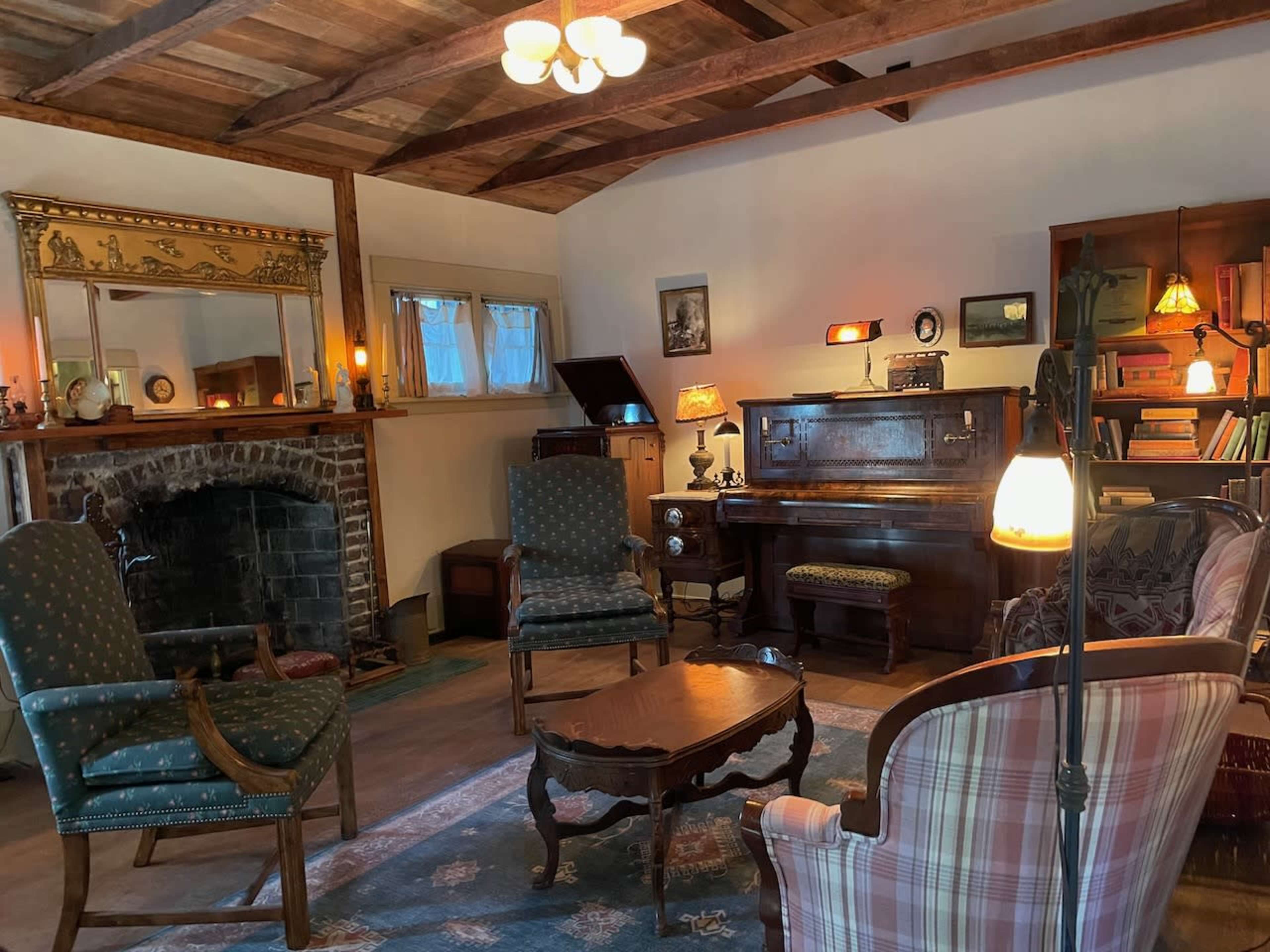 The image shows a vintage living room featuring a piano, two upholstered chairs, a wooden coffee table, and a fireplace, with warm lighting and wooden beams on the ceiling.