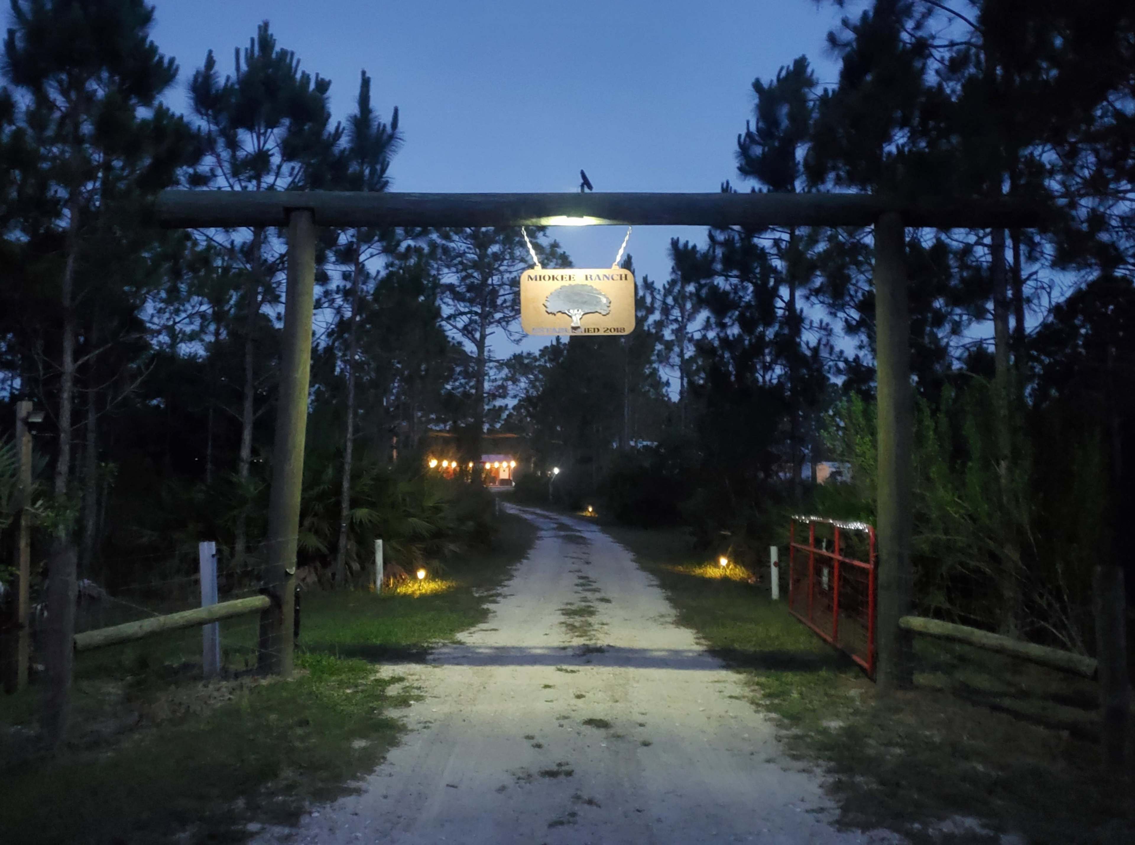 A wooden sign displaying "Morris Ranch" hangs above a gravel road lined with trees and lights at dusk.