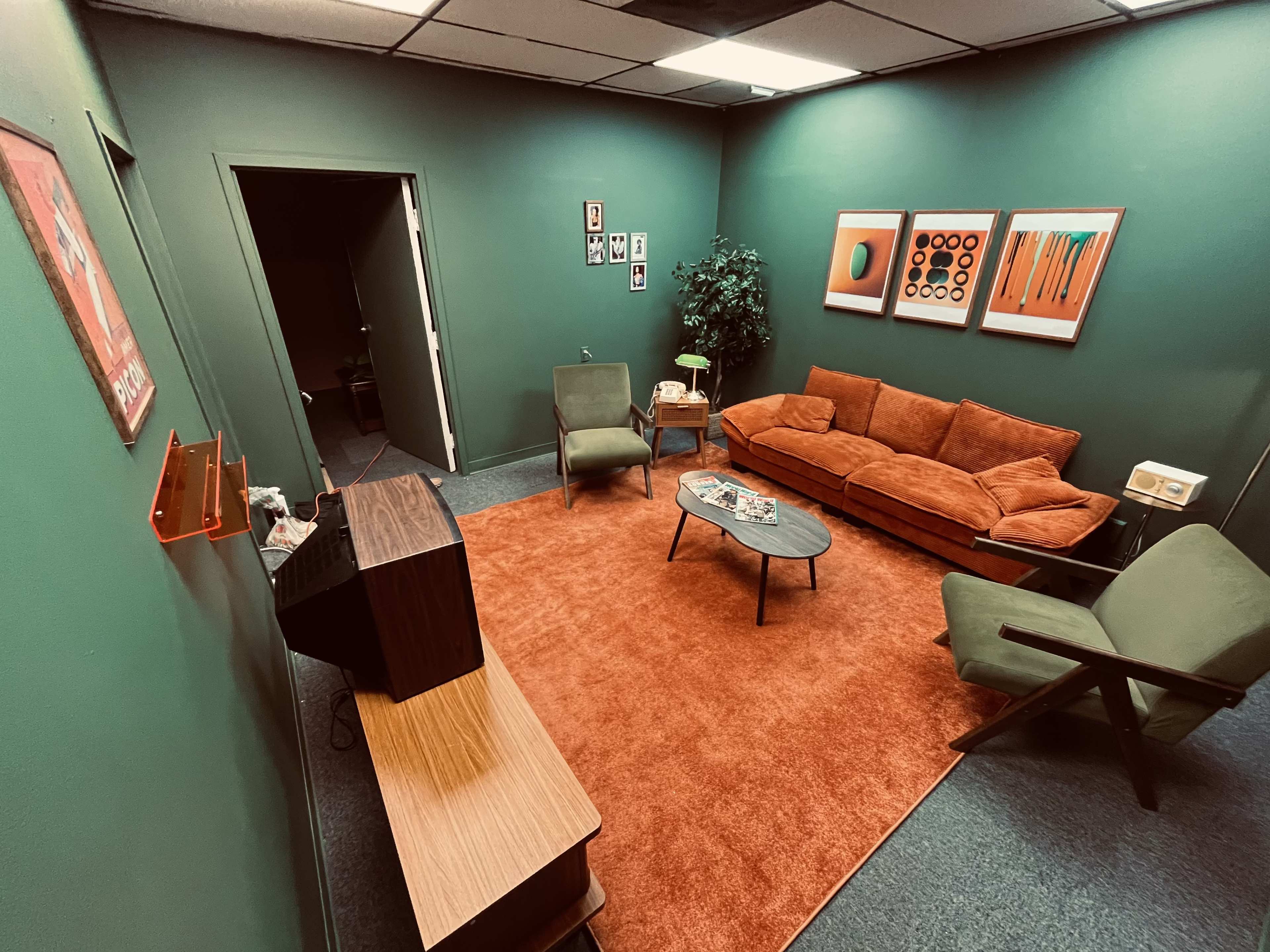 The image shows a cozy room with green walls, featuring an orange couch, two armchairs, a glass coffee table, and a television unit, all arranged on an orange rug.