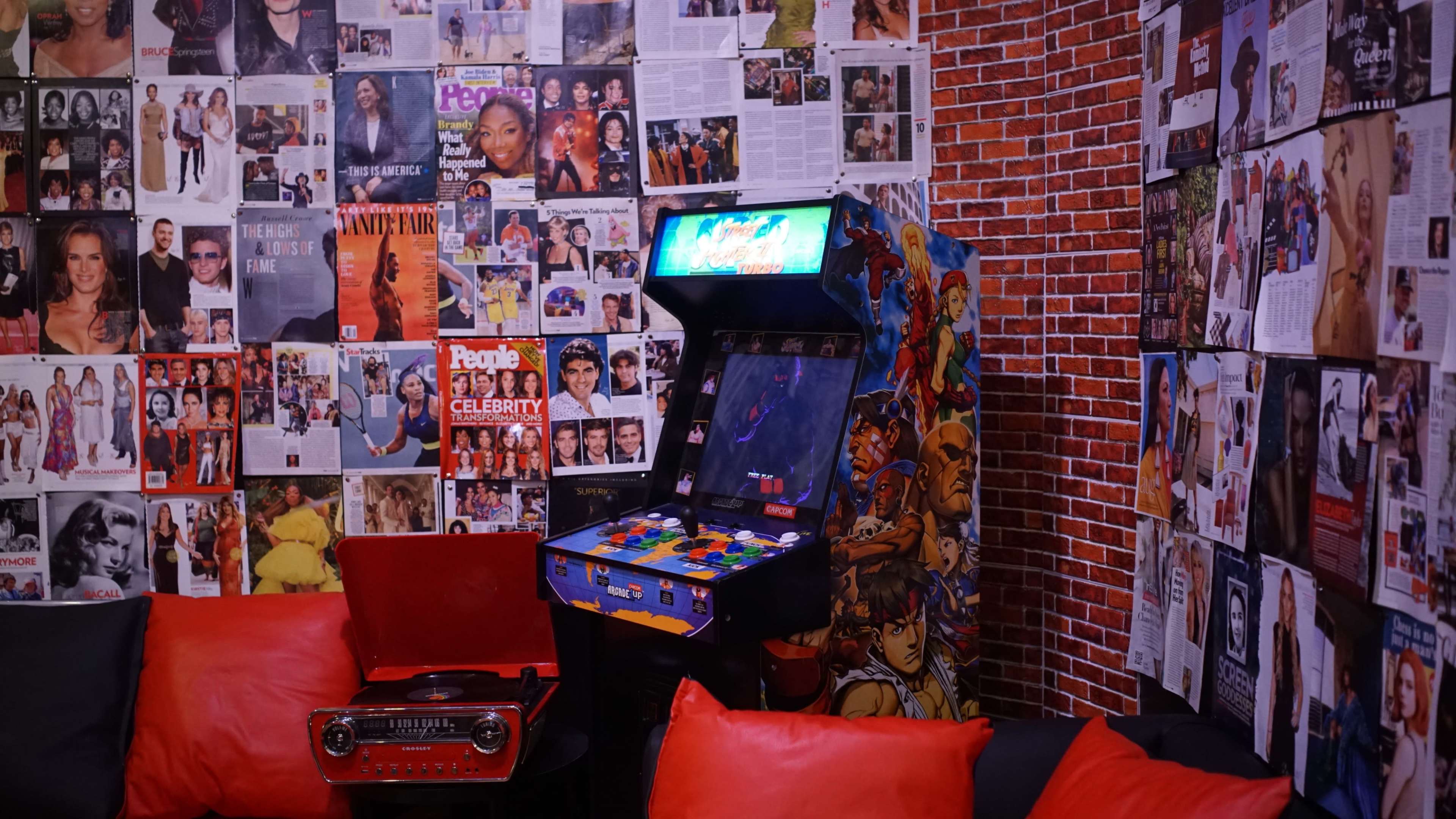 The scene features a cozy room with a video arcade machine, red couches, and walls adorned with various celebrity magazines and posters.