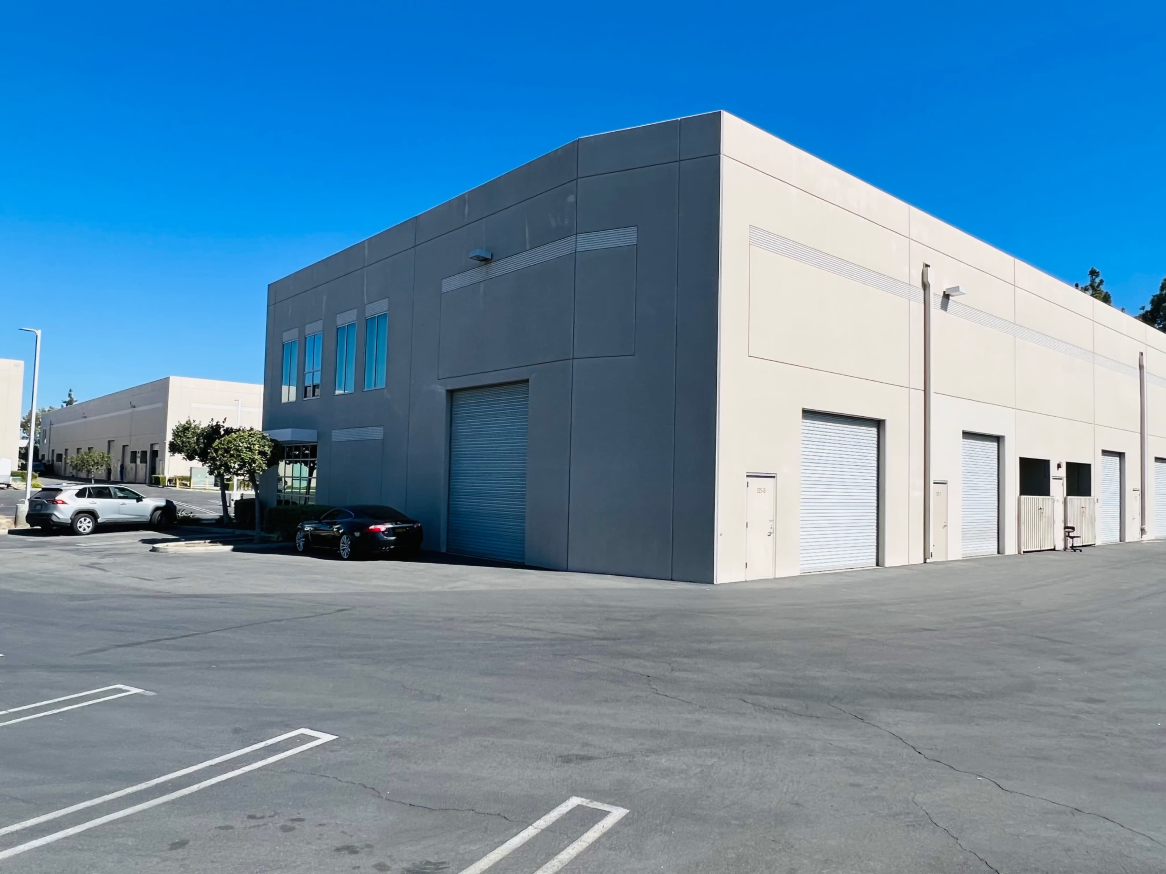 A light-colored industrial building with large roll-up doors sits in a parking lot under a clear blue sky.
