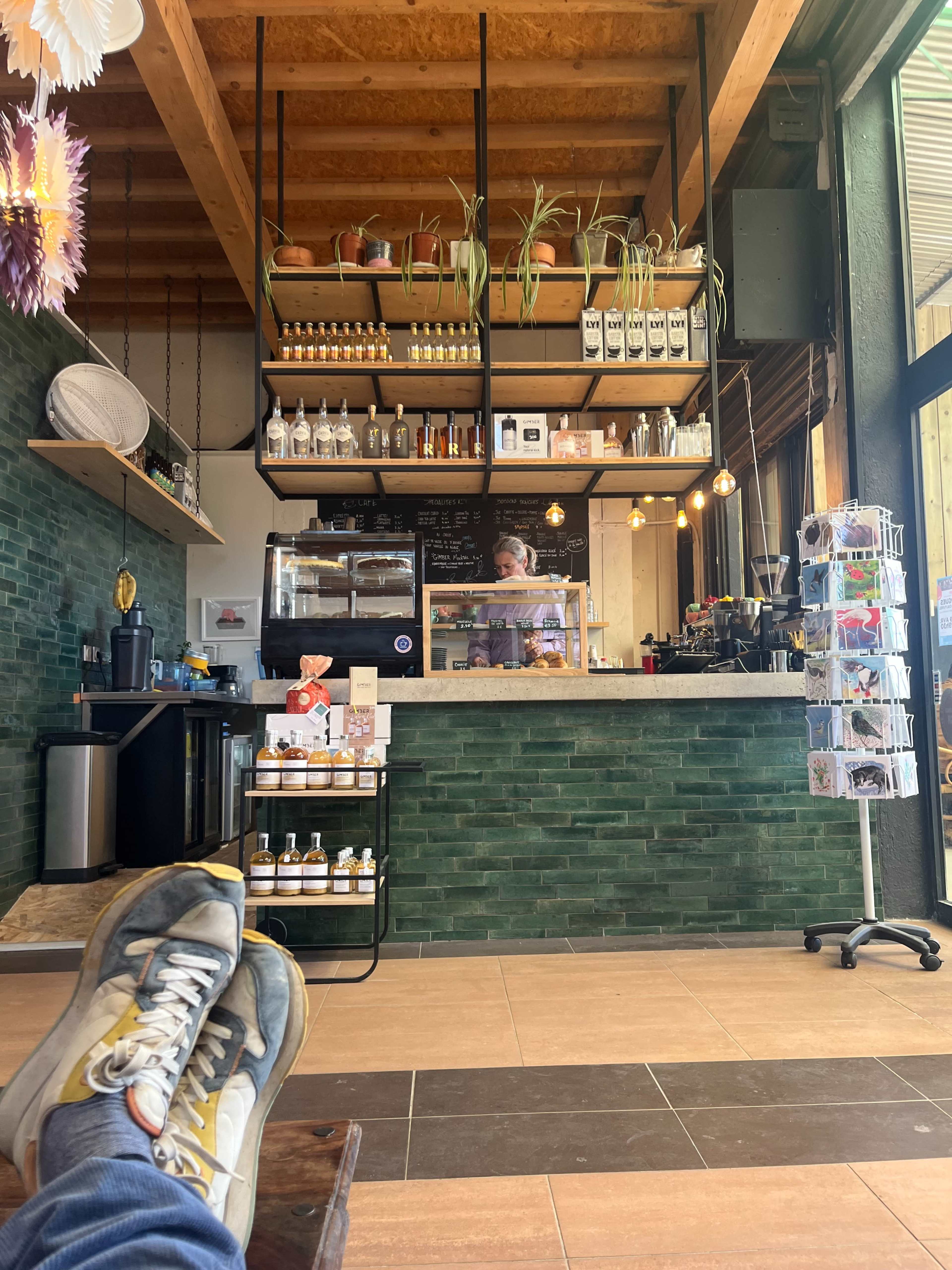 A coffee shop interior features a service counter with a barista, shelves of drinks, and a green tiled wall, while a patron's feet rest on a wooden bench in the foreground.