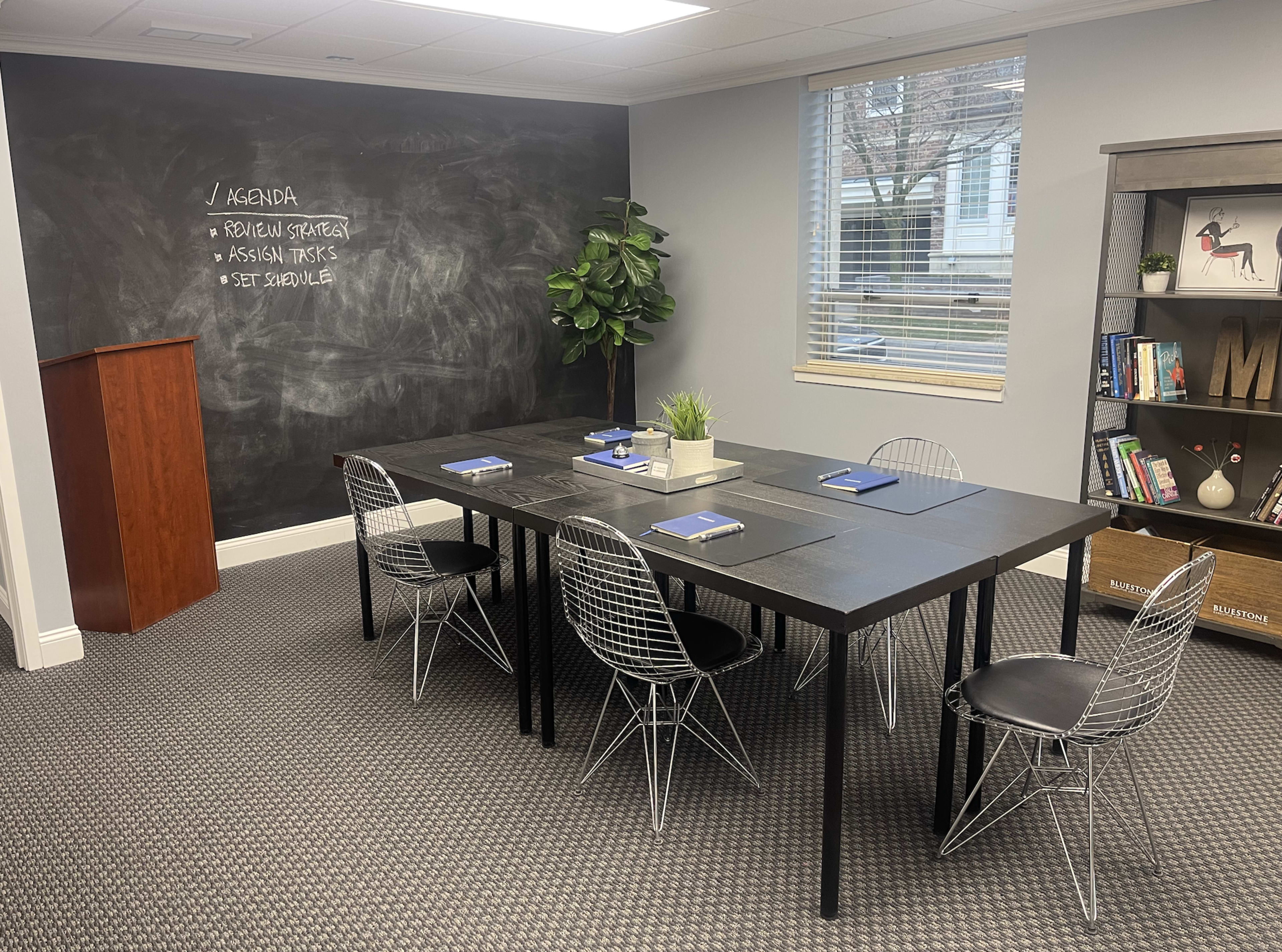 The image shows a conference room with a large black table surrounded by wire chairs, a chalkboard wall listing an agenda, and a bookshelf in one corner.