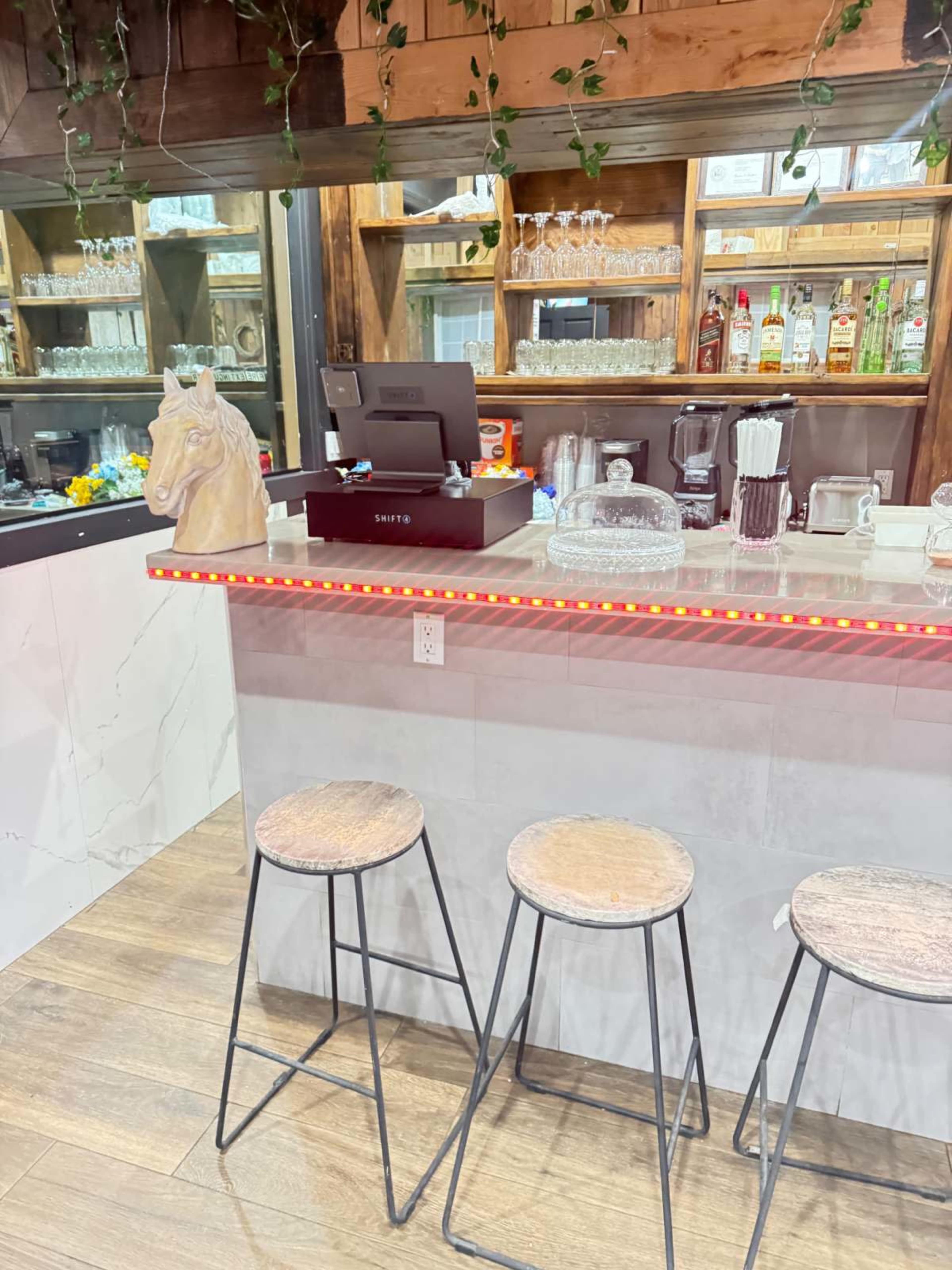 The image shows a bar area featuring three wooden stools, a counter with a cash register, and shelves displaying various bottles and glassware.