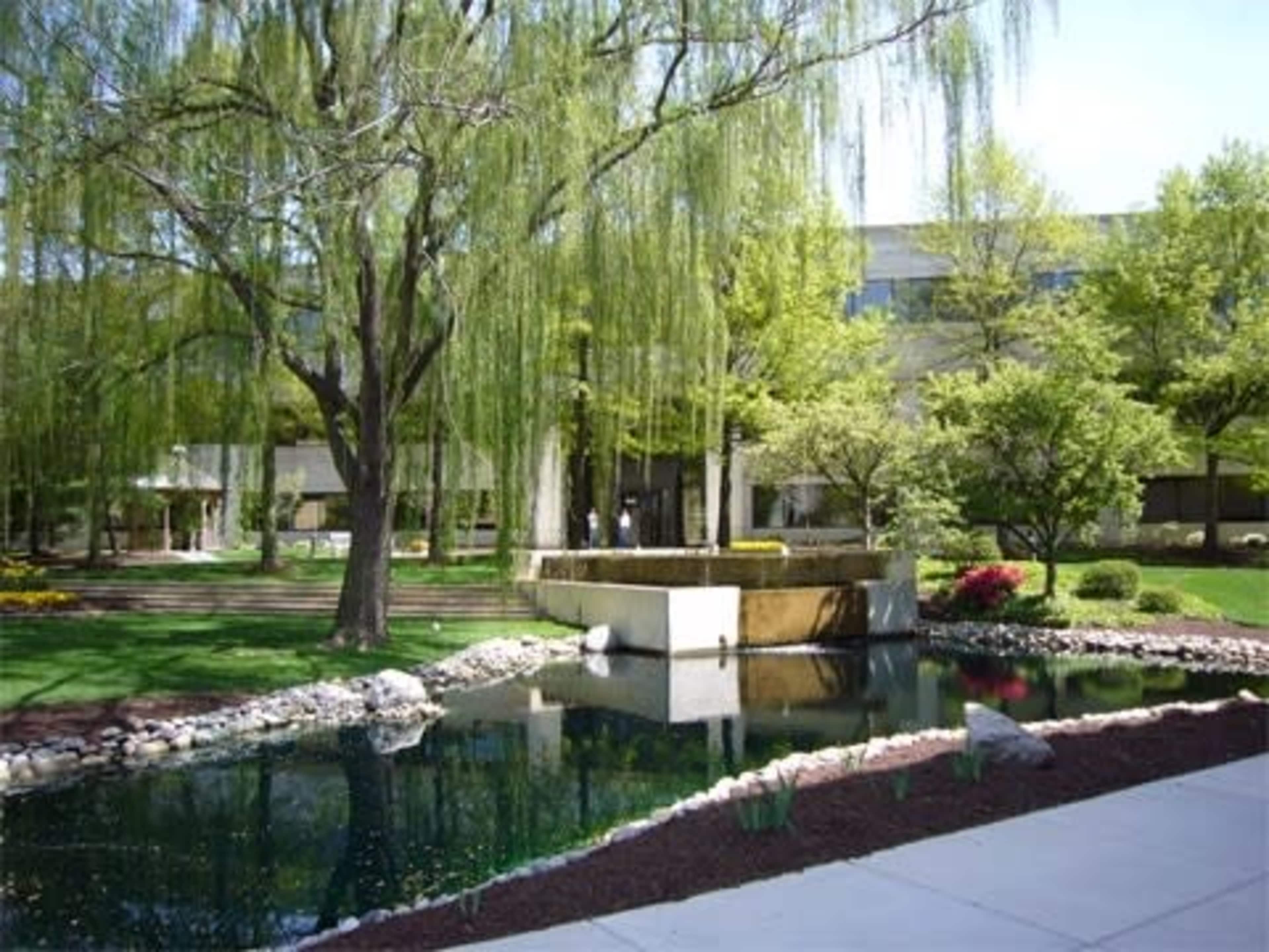 The image shows a landscaped garden with a pond, a stone fountain, and trees, set against a building in the background.