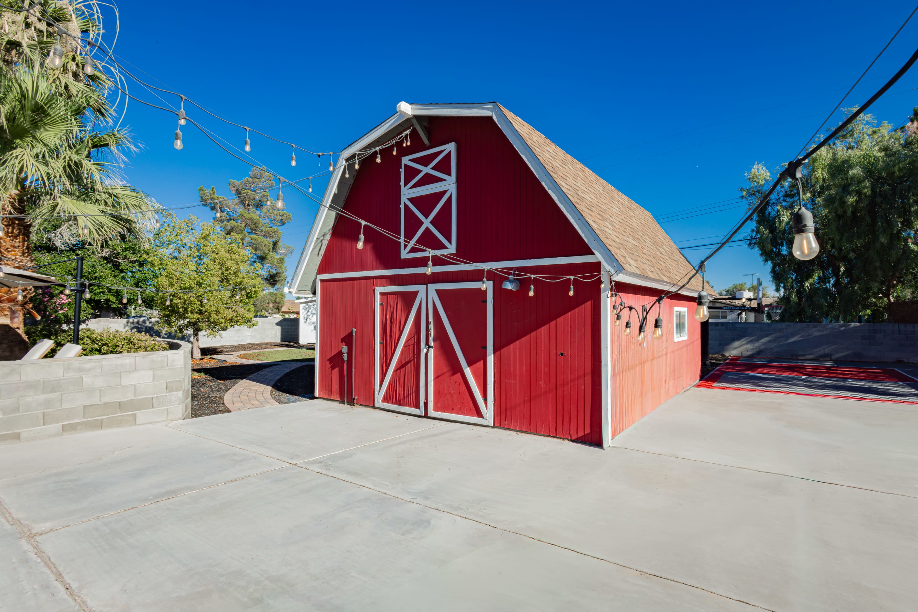 The image features a red barn with a peaked roof, situated on a concrete surface surrounded by greenery and strung lights.