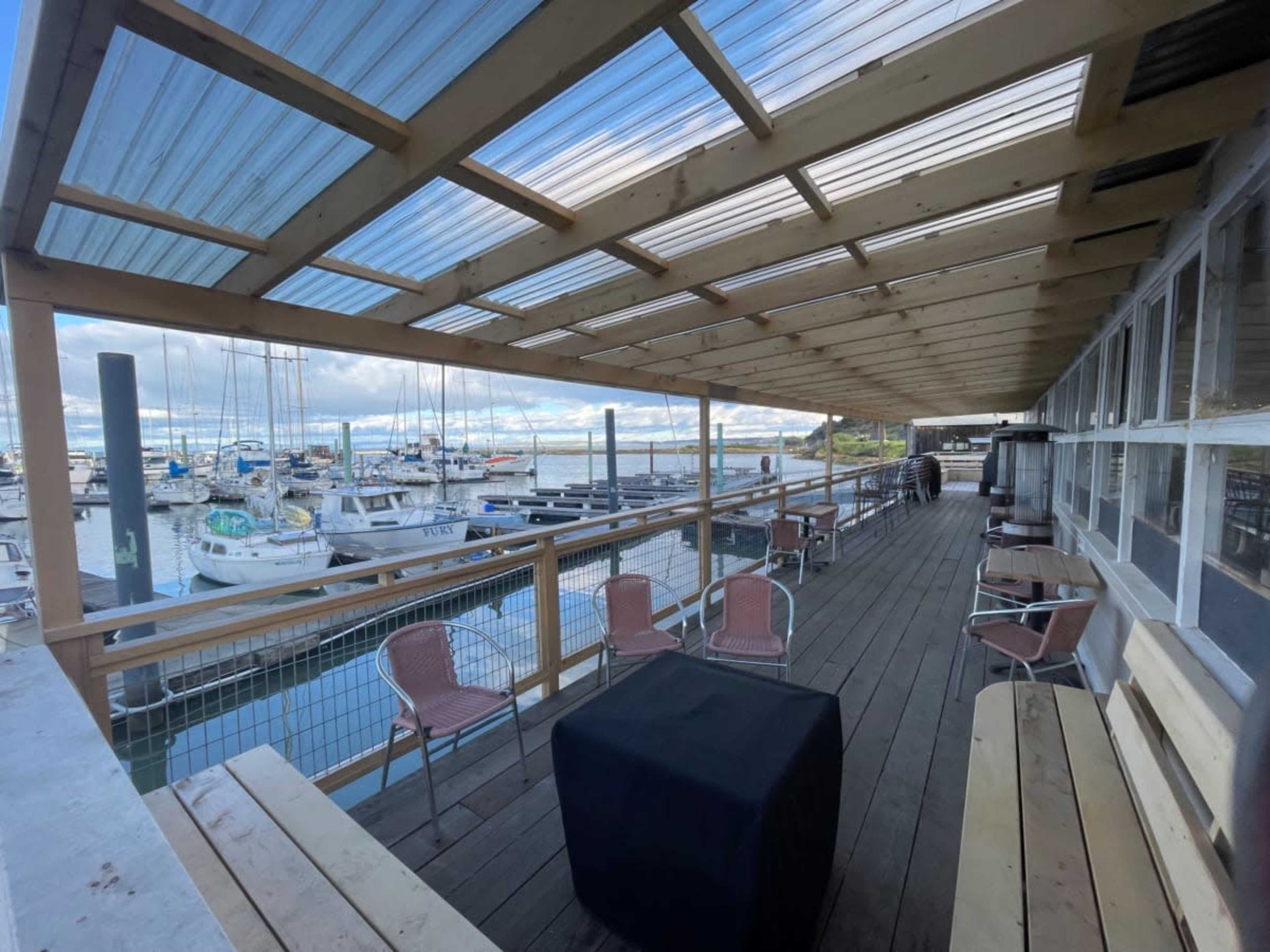 A wooden deck overlooking a marina filled with boats, featuring several seating areas and a partially covered roof.