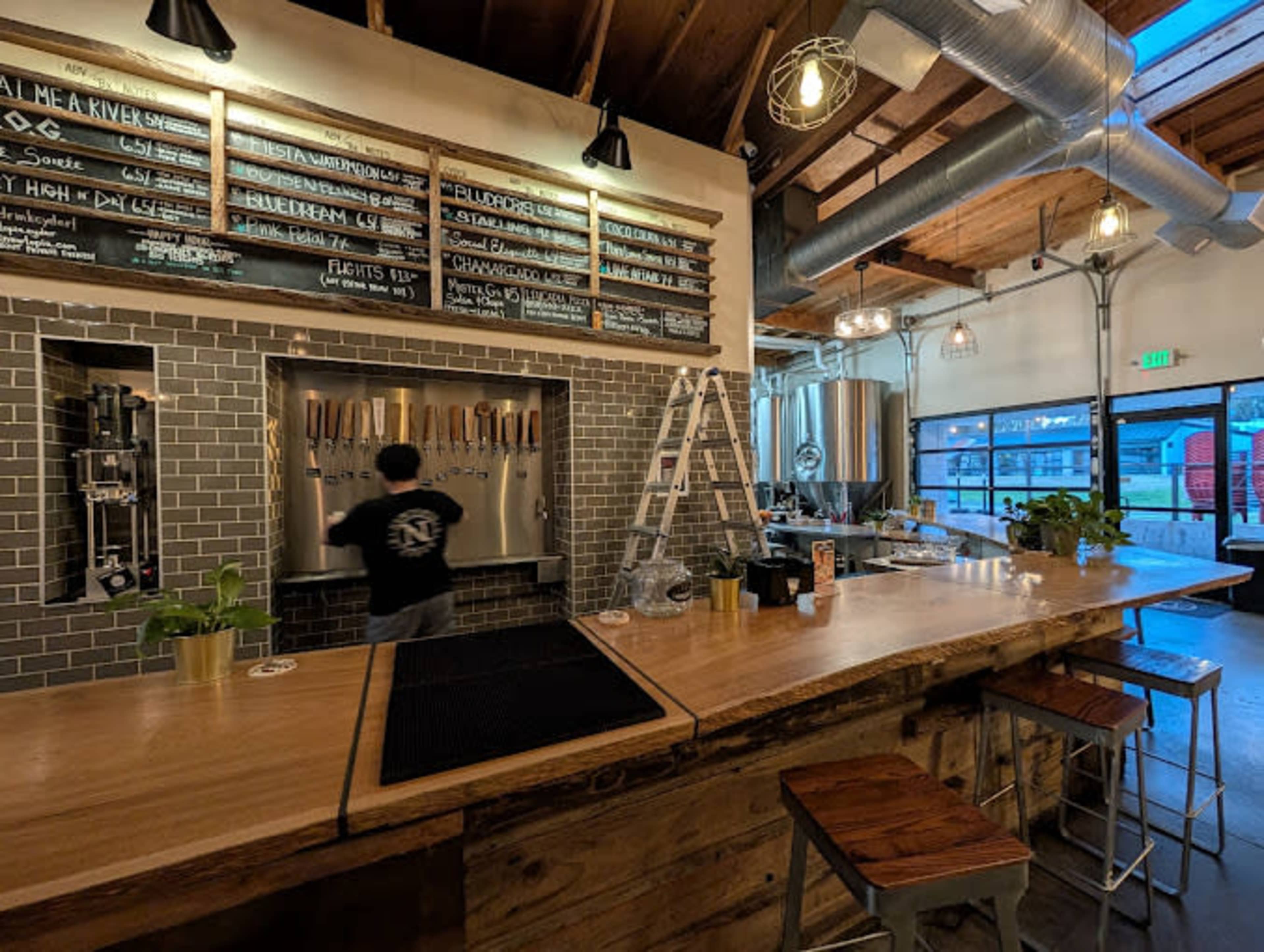 A person is serving drinks at a wooden bar with multiple taps, while a chalkboard menu hangs above, listing various beverages.