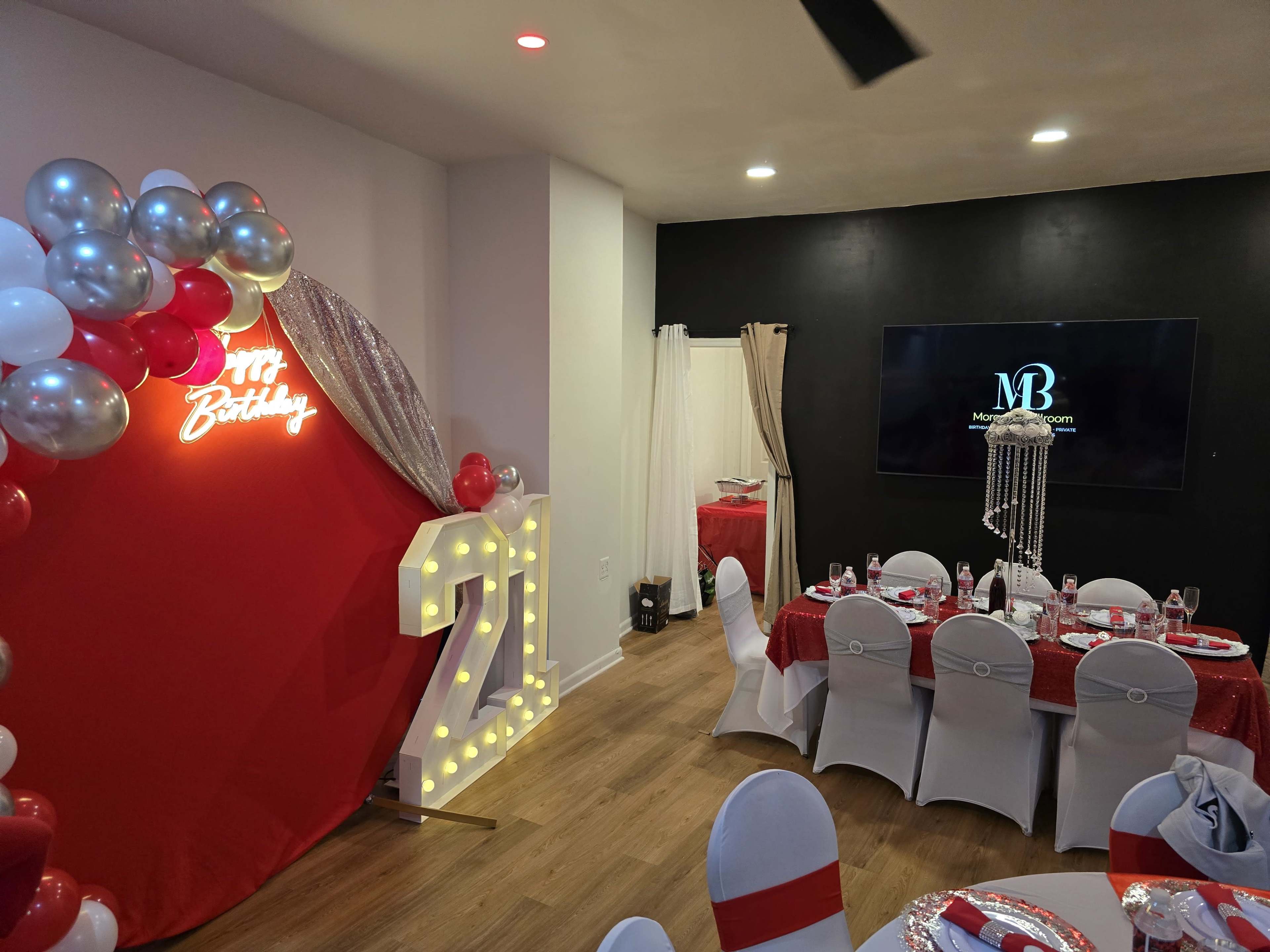 The image shows a festive birthday party setup with a large illuminated sign reading "21," red and silver decorations, and elegantly arranged dining tables.