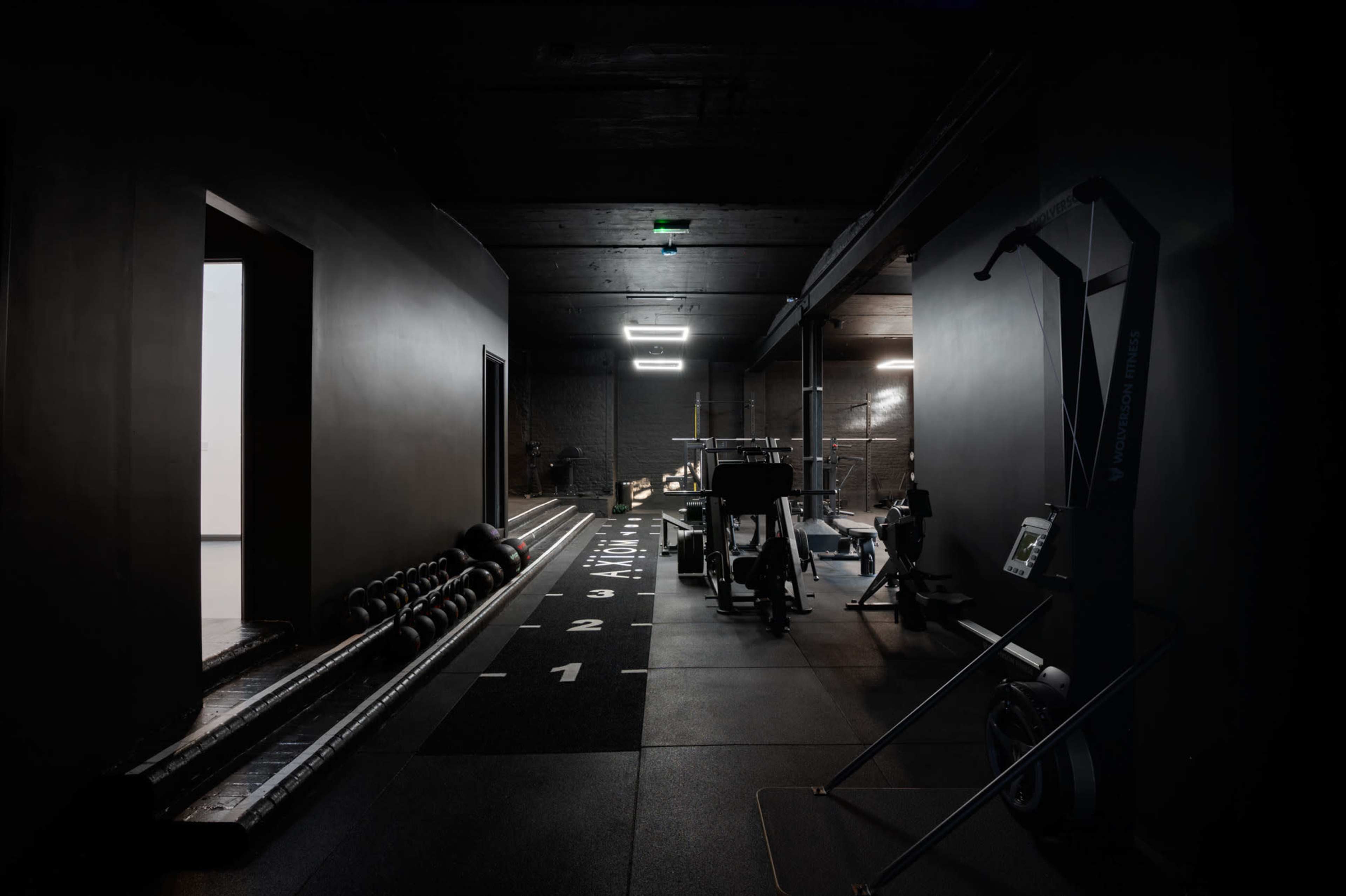 The image shows a dimly lit gym interior featuring various workout equipment, including weights and exercise machines, along a black-floored corridor.