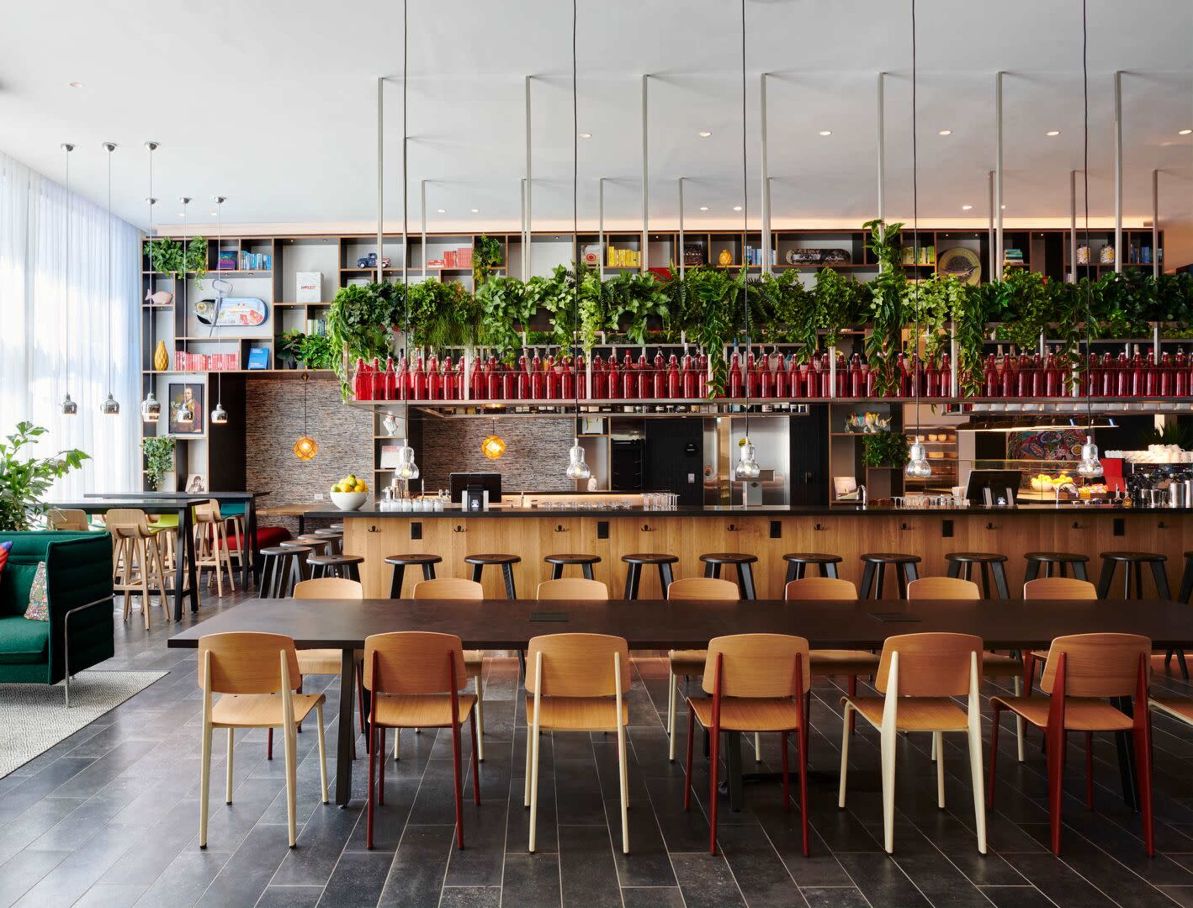 A modern café features a long wooden table surrounded by multicolored chairs, with greenery and shelves filled with decorative items above the bar area.