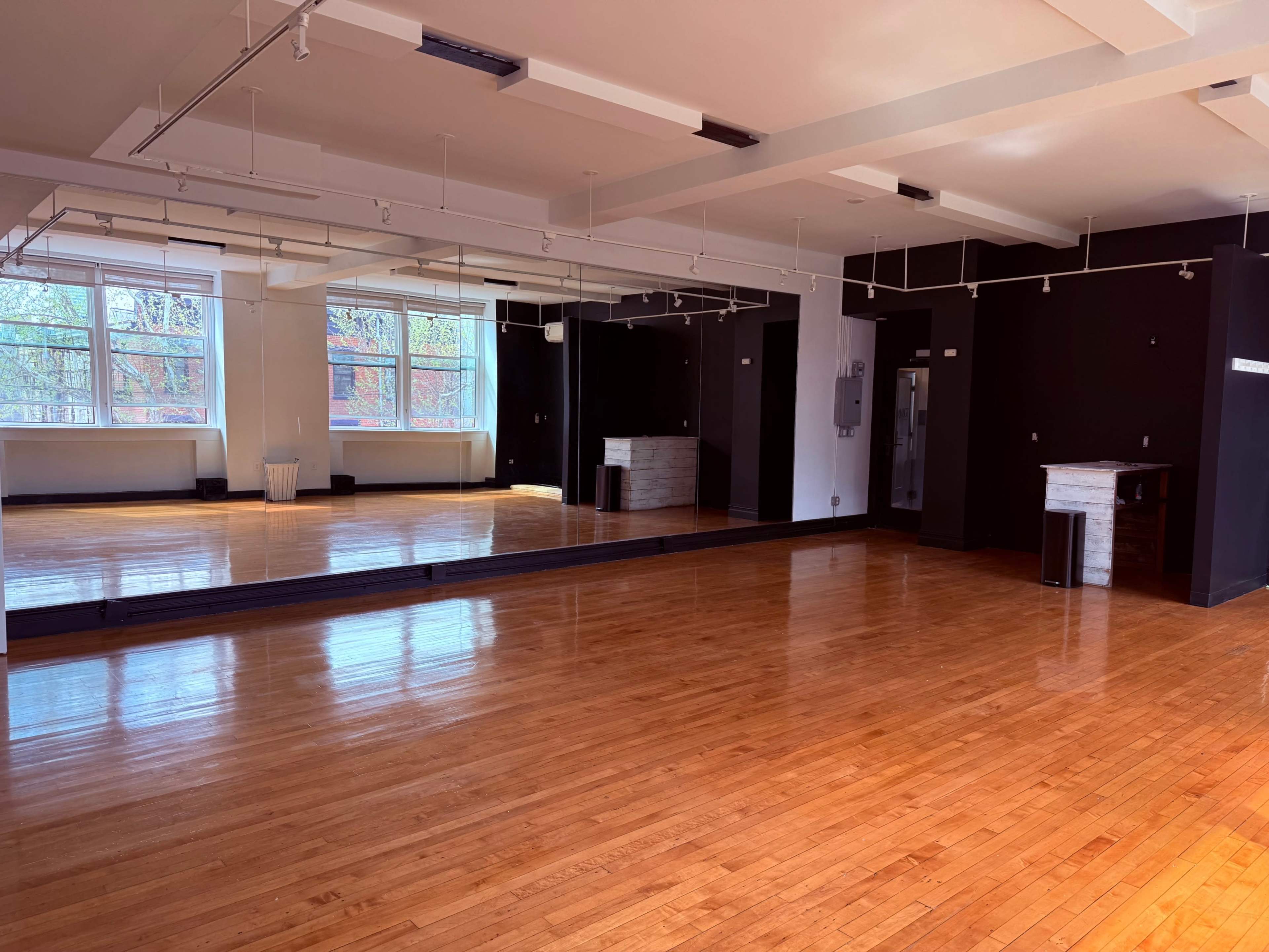The image shows an empty studio space with polished wooden floors, large mirrors on one wall, and windows letting in natural light.