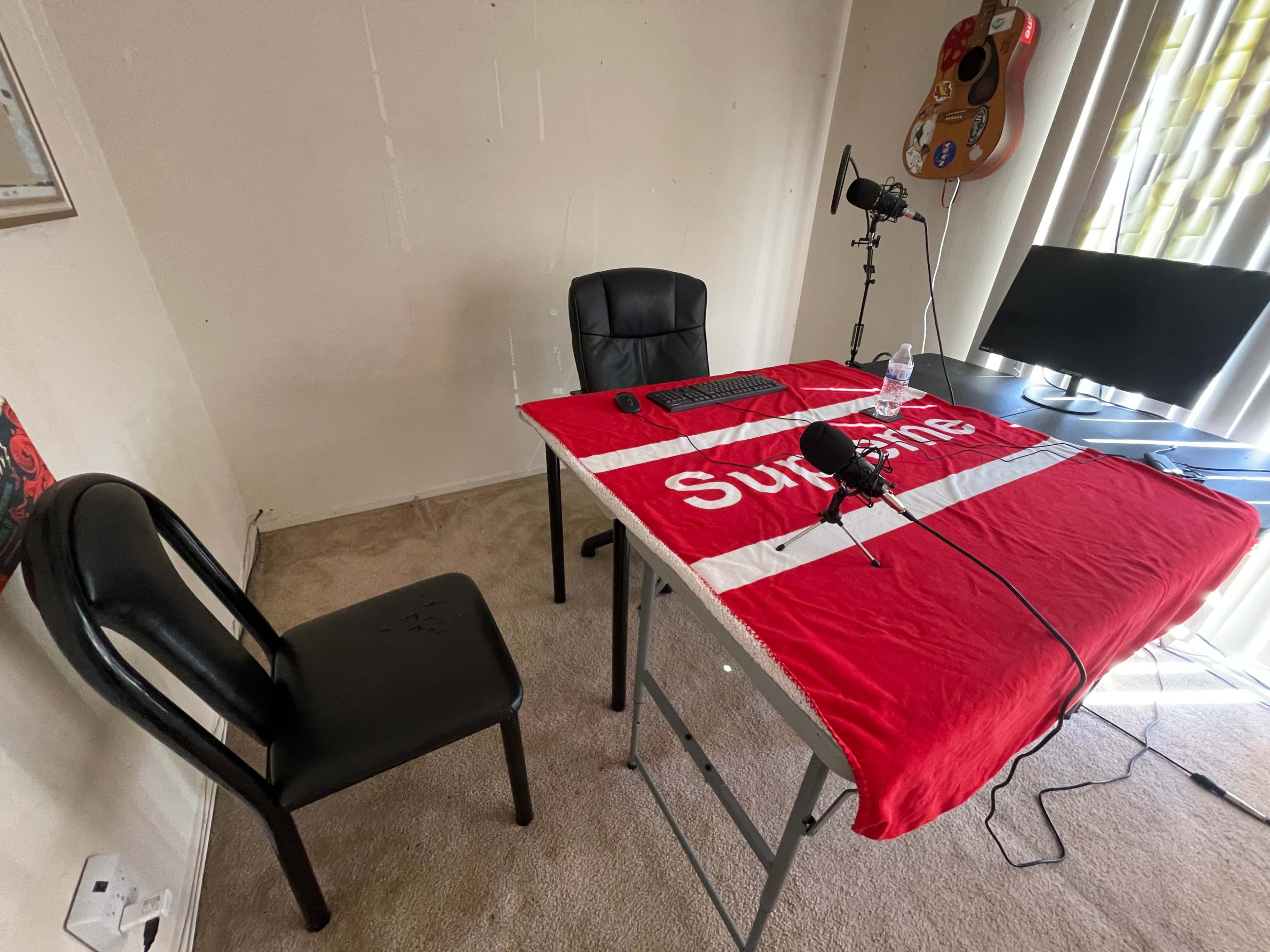 A desk covered with a red flag and equipped with a microphone, a keyboard, and a water bottle is set up in a room with a chair and a guitar hanging on the wall.