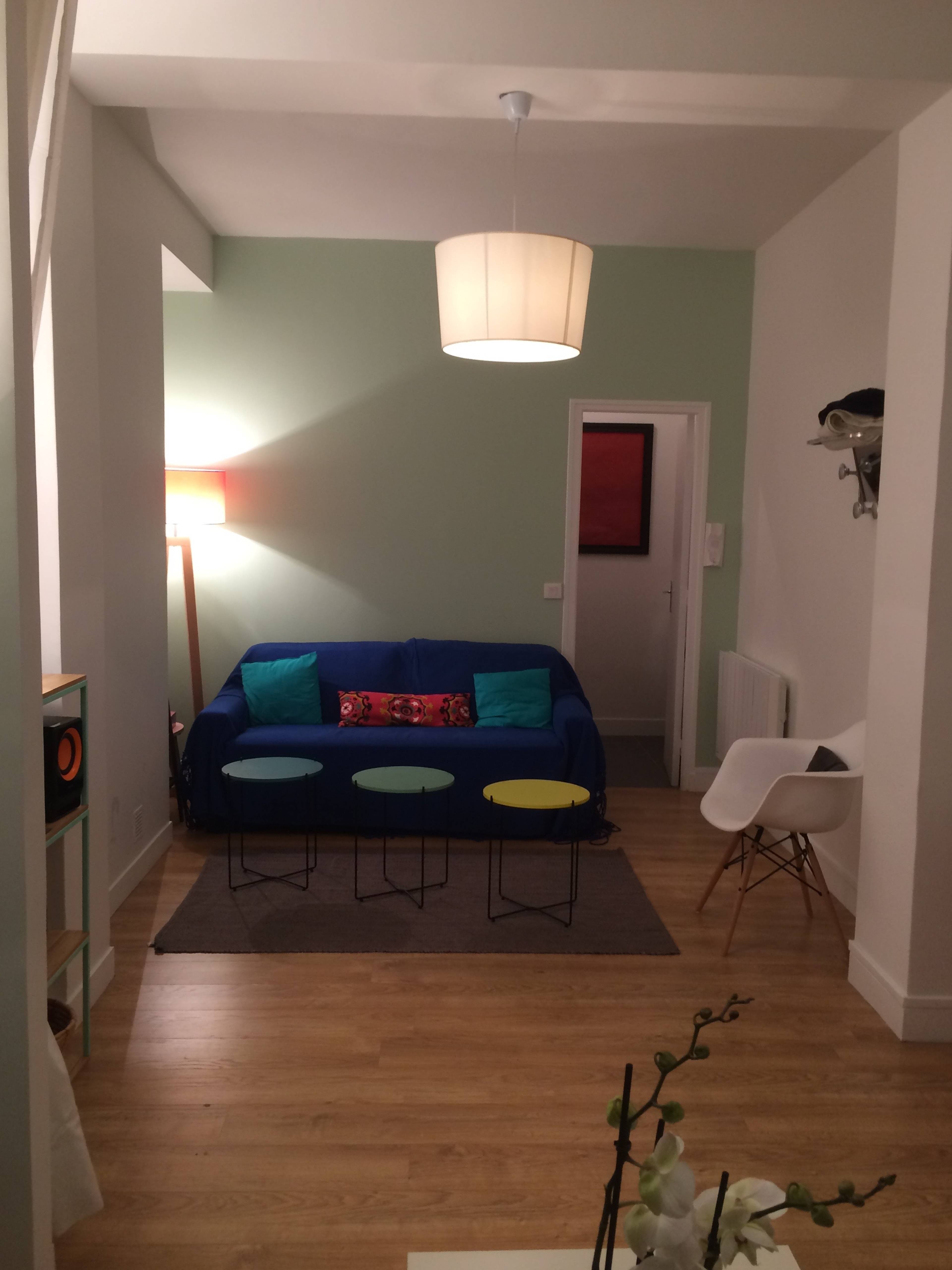 The image depicts a living room with a blue sofa, three colorful stools, a lamp, and a white chair, complemented by light wood flooring.